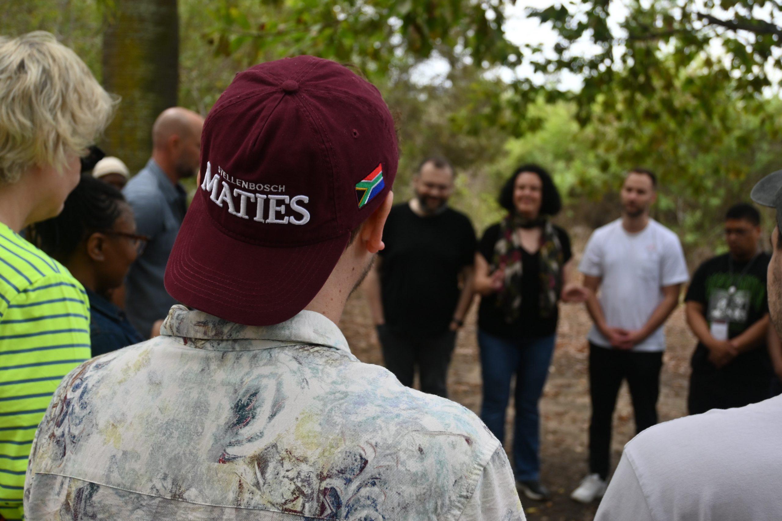 Eine Gruppe von Menschen steht im Freien im Kreis, einer trägt eine kastanienbraune Stellenbosch-Maties-Mütze mit einer kleinen südafrikanischen Flaggennadel. Im Hintergrund sind Bäume und Grünflächen zu sehen.