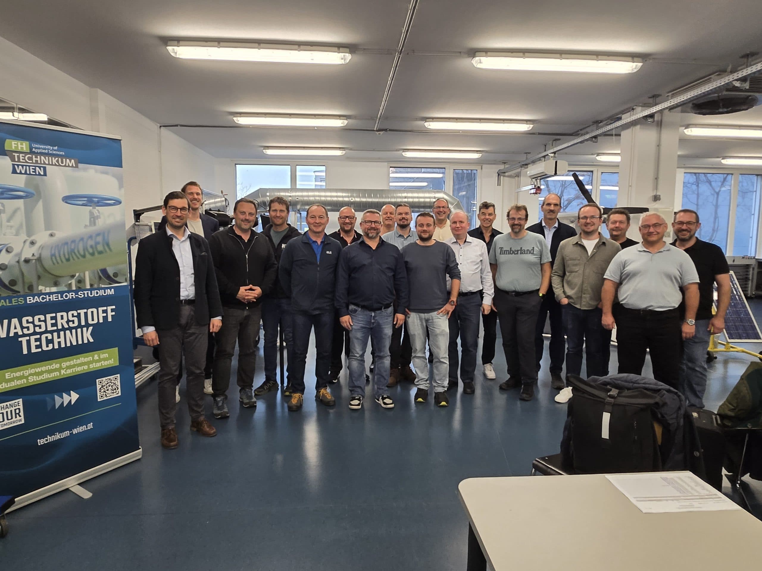 Eine Gruppe von etwa zwanzig Personen posiert und lächelt für ein Foto in einem hellen Klassenzimmer, das neben einem blauen Banner steht, das für den Studiengang Wasserstofftechnologie an der FH Technikum Wien wirbt.