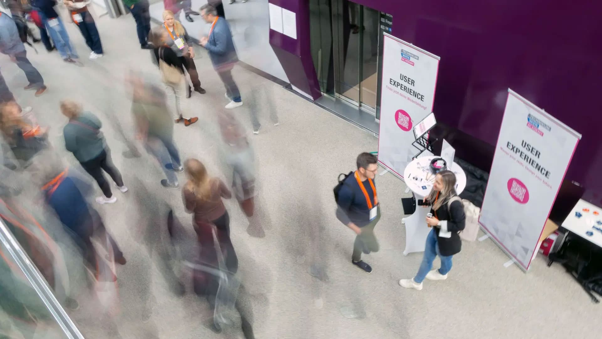 Eine unscharfe Draufsicht auf Menschen, die sich in einem belebten Innenflur einer Veranstaltung bewegen und unterhalten, mit zwei Personen, die in der Nähe eines User-Experience-Standes an einer lila Wand stehen.