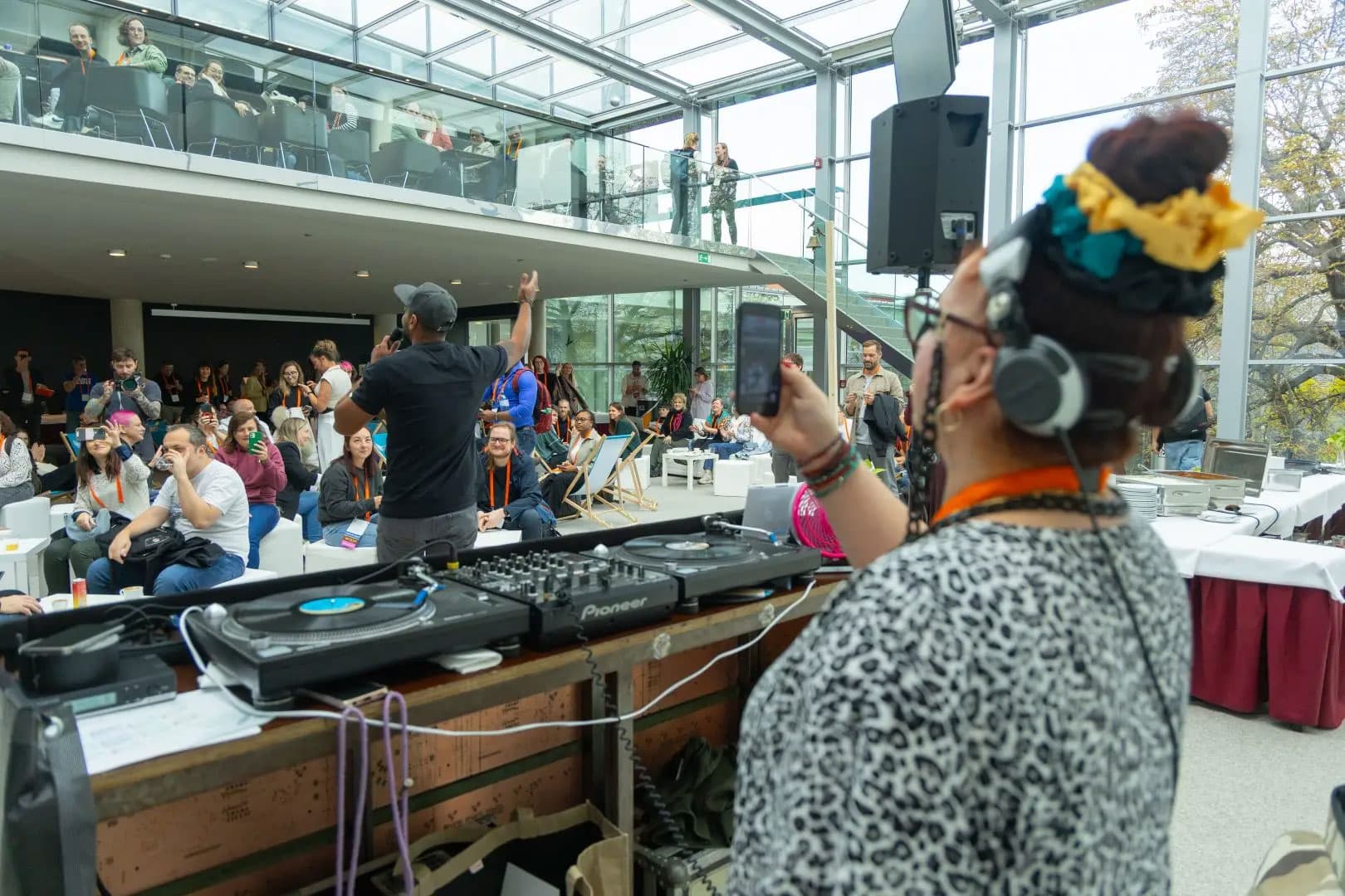 Ein DJ mit Kopfhörern legt bei einer Indoor-Veranstaltung Musik auf. Die Leute sitzen und stehen, einige heben die Hände. Der Veranstaltungsort hat große Fenster und ein Zwischengeschoss mit mehr Teilnehmern.