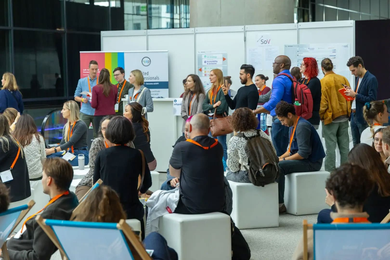 Eine bunt gemischte Gruppe von Menschen mit Konferenzausweisen sitzt und steht, hört zu und unterhält sich in einem modernen Veranstaltungsraum mit Informationsständen und Postern im Hintergrund.