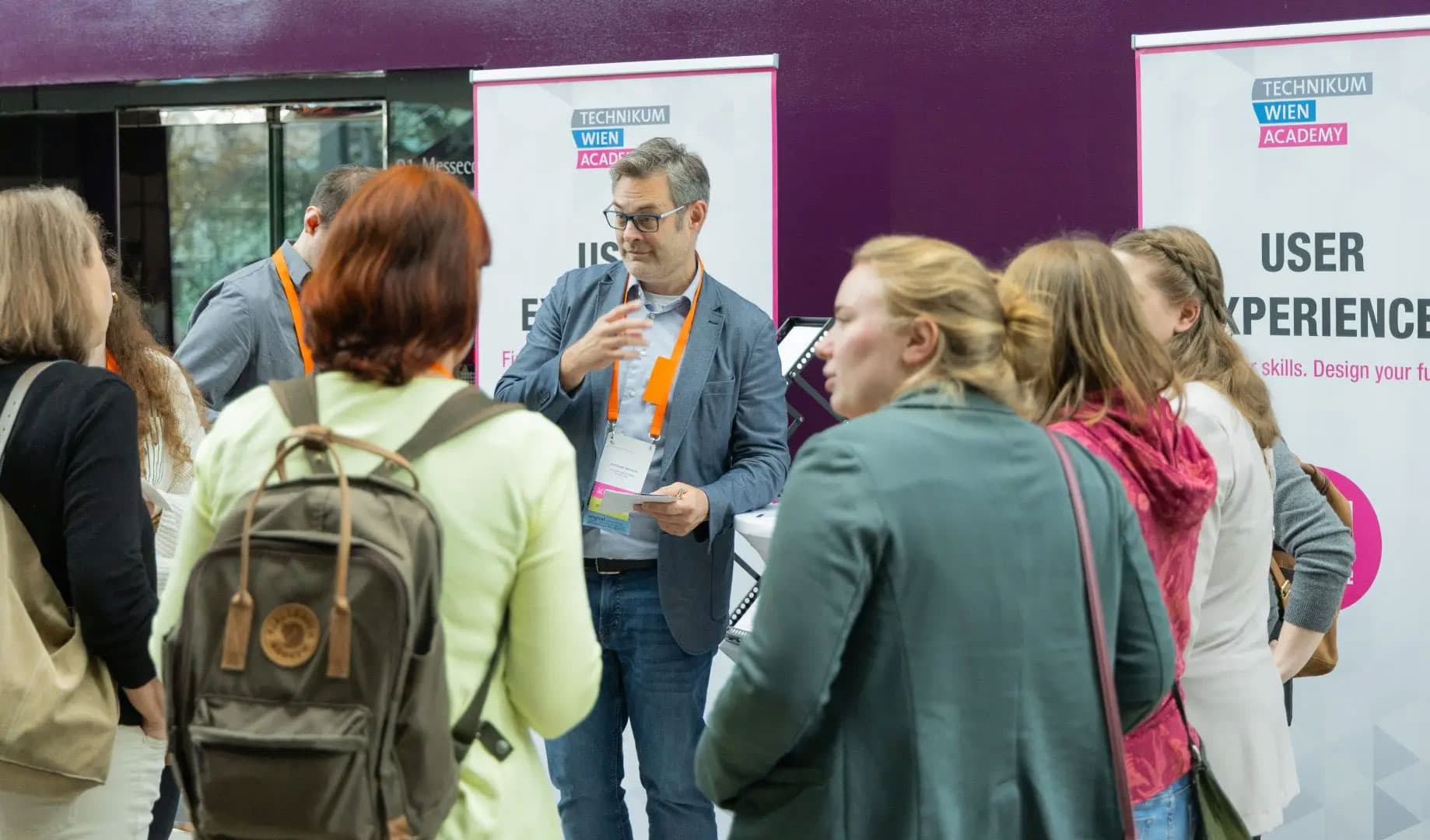 Eine Gruppe von Menschen steht im Halbkreis um einen Mann, der Papiere in der Hand hält und bei einer Veranstaltung der Technikum Wien Academy spricht. Im Hintergrund sind Banner zum Thema User Experience zu sehen.