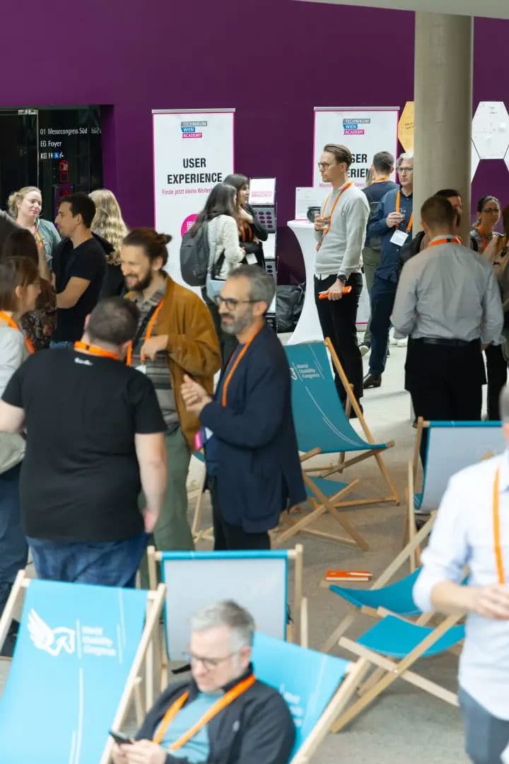 Eine Gruppe von Personen mit orangefarbenen Schlüsselbändern unterhält sich bei einer Veranstaltung in einer Halle. Einige stehen und unterhalten sich in der Nähe von User Experience-Bannern, während andere auf blauen Liegestühlen im Vordergrund sitzen. Die Wand im Hintergrund ist lila.