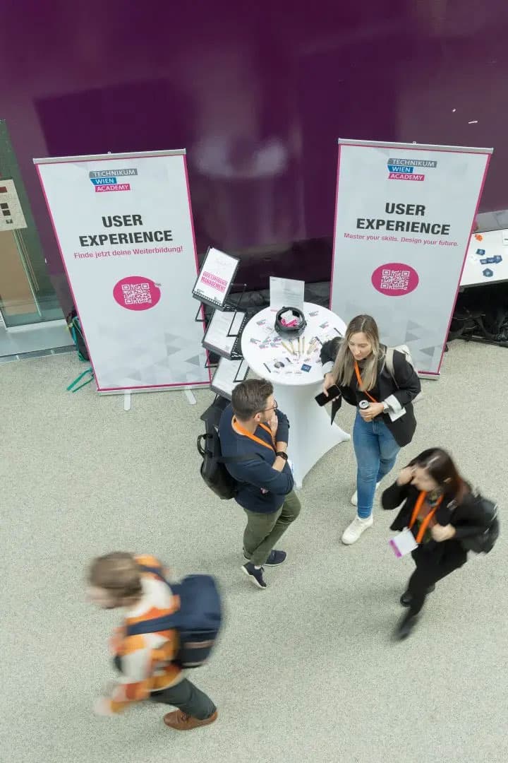 Draufsicht auf drei Personen, die sich an einem runden Tisch mit Broschüren auf einer Veranstaltung über Benutzererfahrung unterhalten. Hinter ihnen zwei große Banner mit der Aufschrift USER EXPERIENCE vor einer lila Wand.