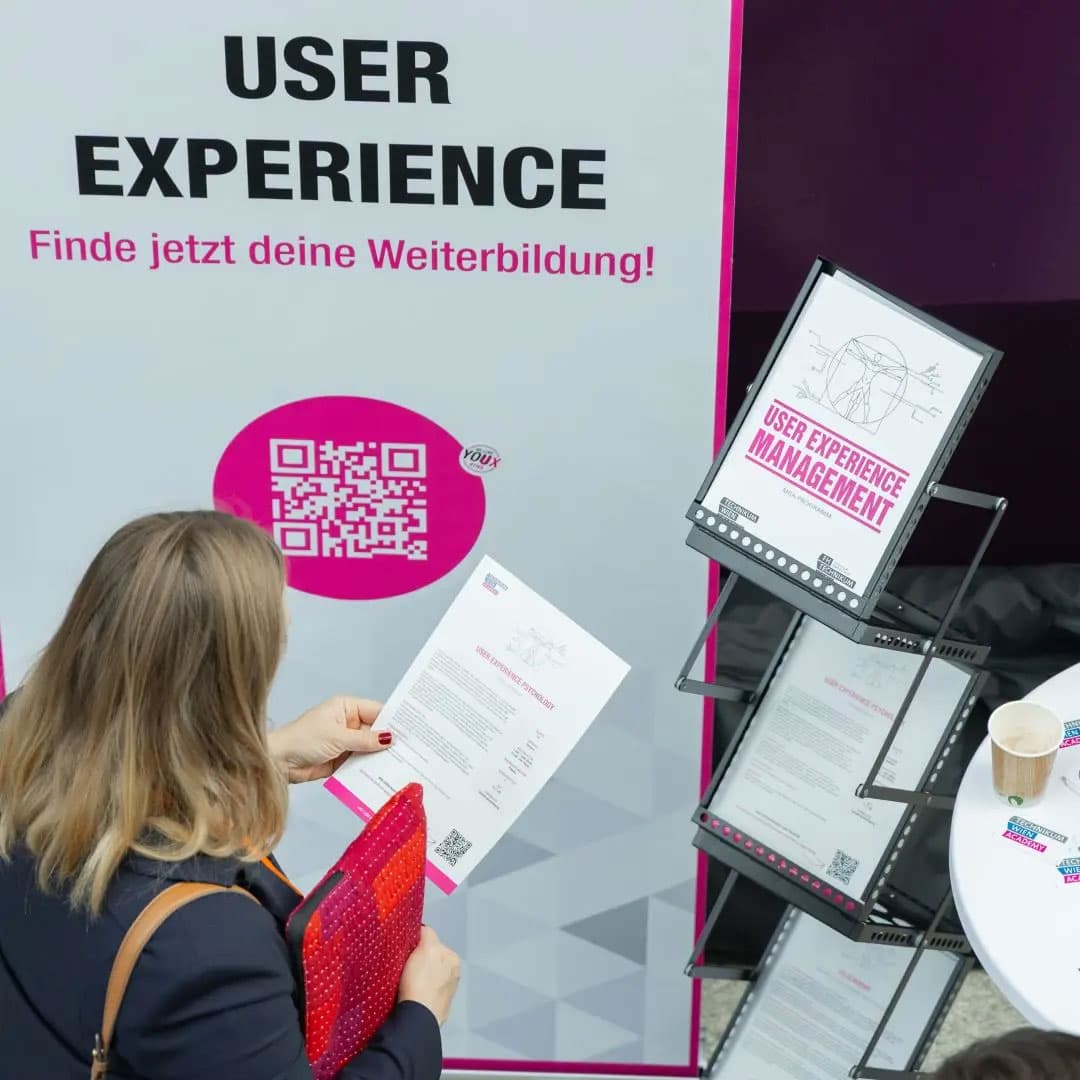 Eine Frau mit einer Zeitung in der Hand steht vor einem Display zum Thema User Experience mit einem QR-Code, Informationsflyern und einem deutschen Text, der für Weiterbildung wirbt. Eine Kaffeetasse steht auf einem kleinen Tisch in der Nähe.