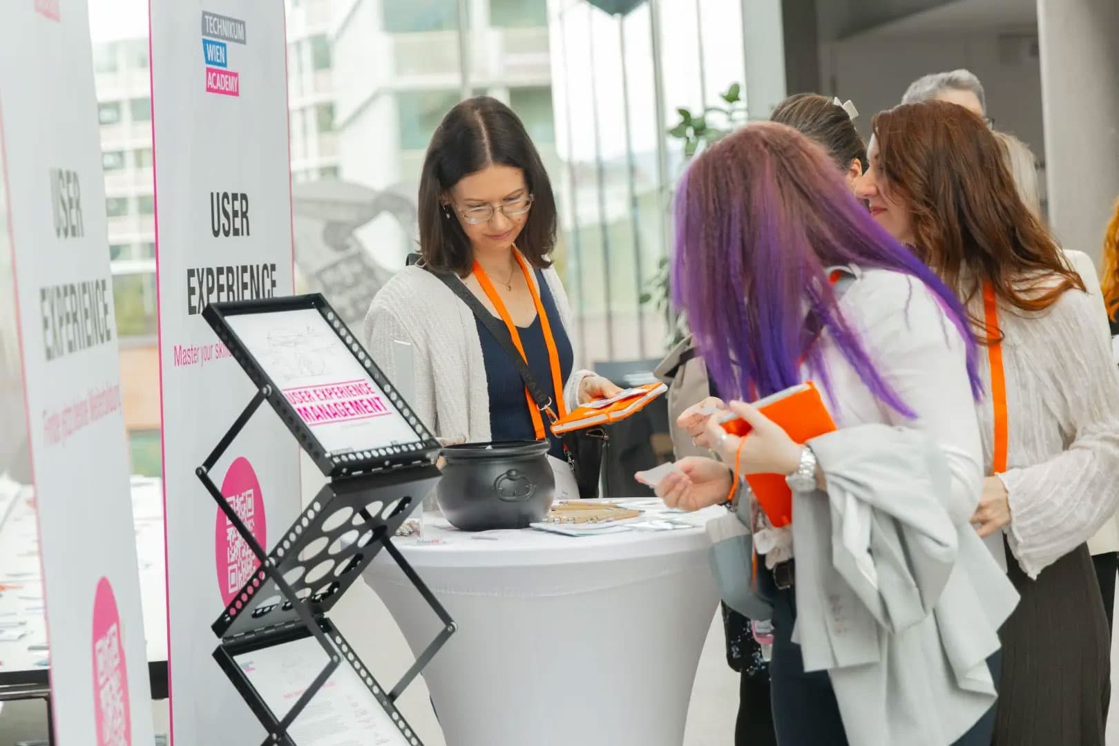 Mehrere Frauen mit Konferenzbändern stehen um einen hohen runden Tisch mit einem Benutzererfahrungsschild, Stiften und Notizbüchern und beschäftigen sich mit den Veranstaltungsmaterialien in einem hellen Innenraum.