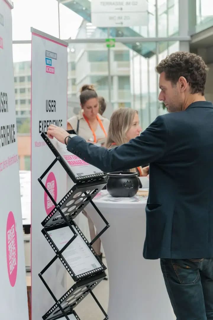 Ein Mann liest an einem Stand für Benutzererfahrungen Broschüren aus einem Regal, während zwei Frauen an einem Tisch in der Nähe in einer modernen, hellen Konferenzumgebung arbeiten.