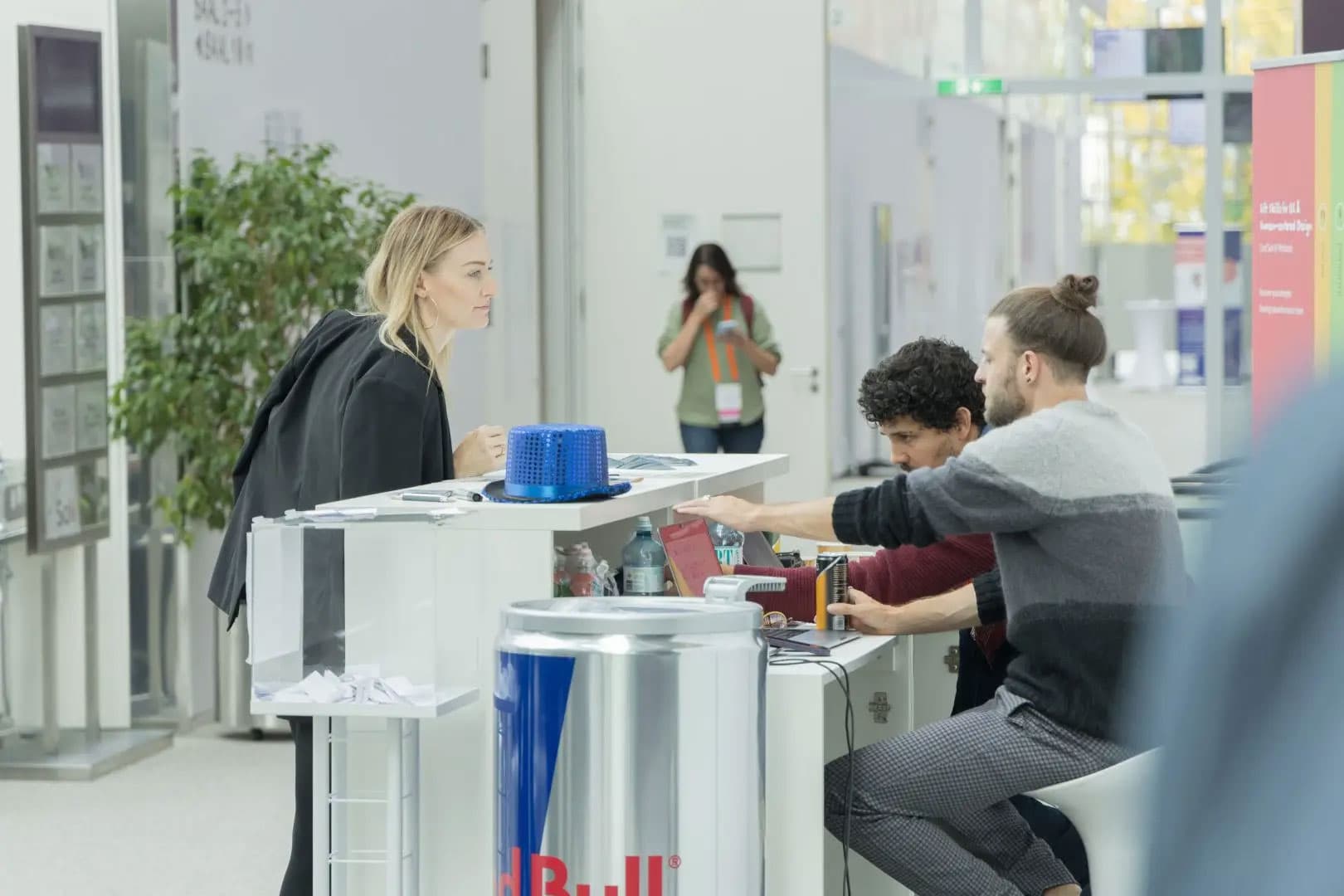 Eine Frau steht an der Rezeption und unterhält sich mit zwei sitzenden Männern in einer modernen, hellen Innenumgebung. Im Hintergrund spricht eine weitere Person am Telefon. Im Vordergrund steht ein Tisch in Form einer Red Bull-Dose.