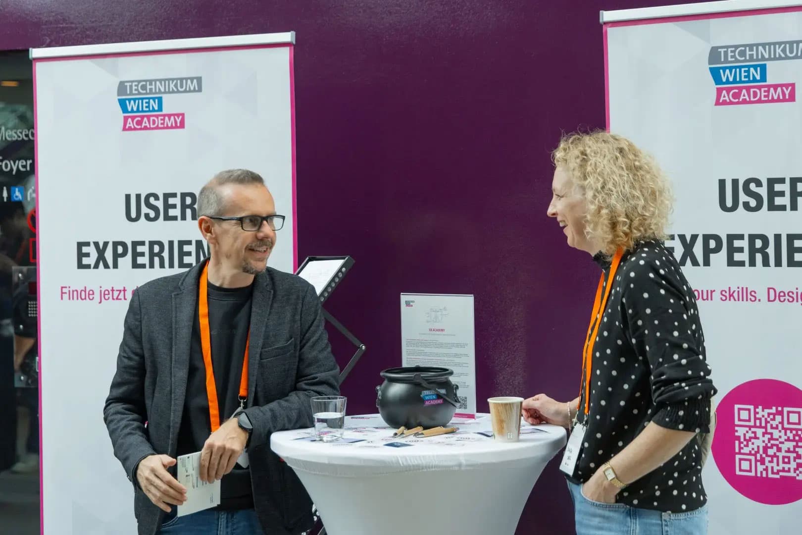 Zwei Personen stehen während einer Veranstaltung an einem runden Tisch und unterhalten sich. Hinter ihnen sind Banner mit der Aufschrift Technikum Wien Academy und User Experience zu sehen. Beide tragen orangefarbene Schlüsselbänder und halten Getränke in der Hand, auf dem Tisch liegen Flyer und Gegenstände.