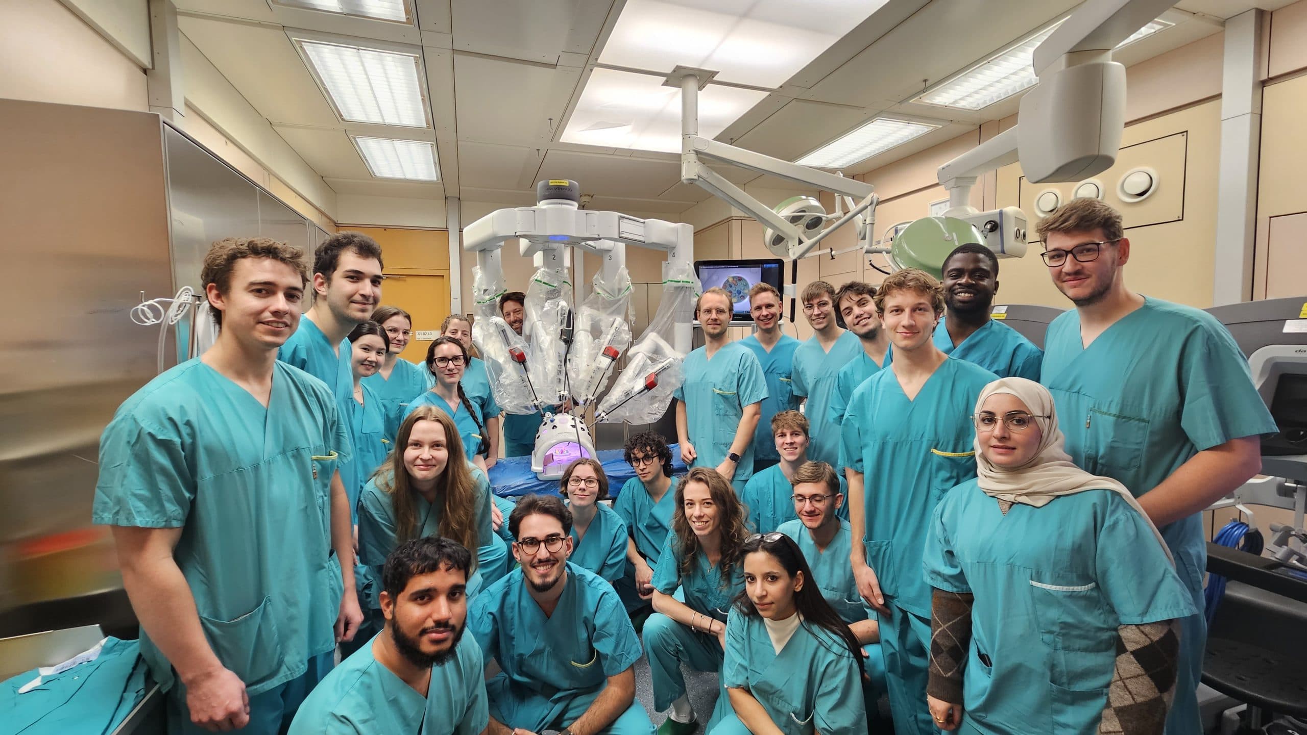 Eine Gruppe von Medizinstudenten in blaugrünen Kitteln posiert zusammen in einem Operationssaal mit medizinischen Geräten und einem chirurgischen Roboter im Hintergrund.