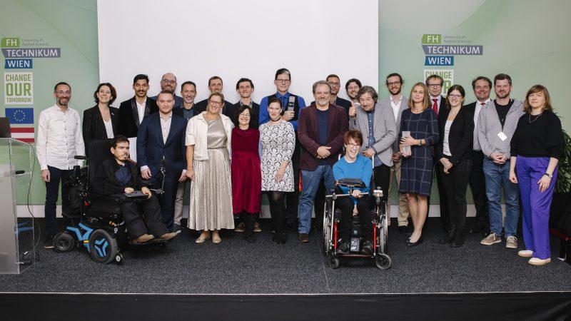 Eine Gruppe von etwa 25 Personen, darunter Männer und Frauen, einige im Rollstuhl, posieren gemeinsam auf einer Bühne vor Transparenten mit der Aufschrift 'FH Technikum Wien' und 'Change Our Tomorrow'.