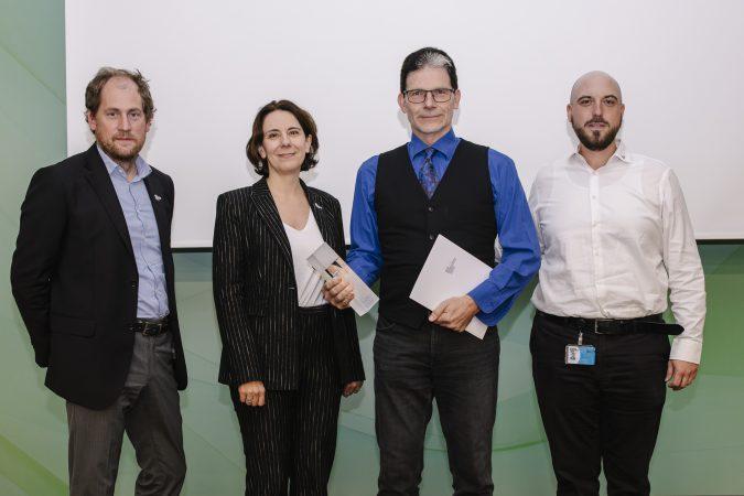 Vier Personen stehen nebeneinander in formeller Kleidung, zwei halten Urkunden oder Auszeichnungen vor einem grün-weißen Hintergrund und posieren für ein Gruppenfoto bei einer Veranstaltung.