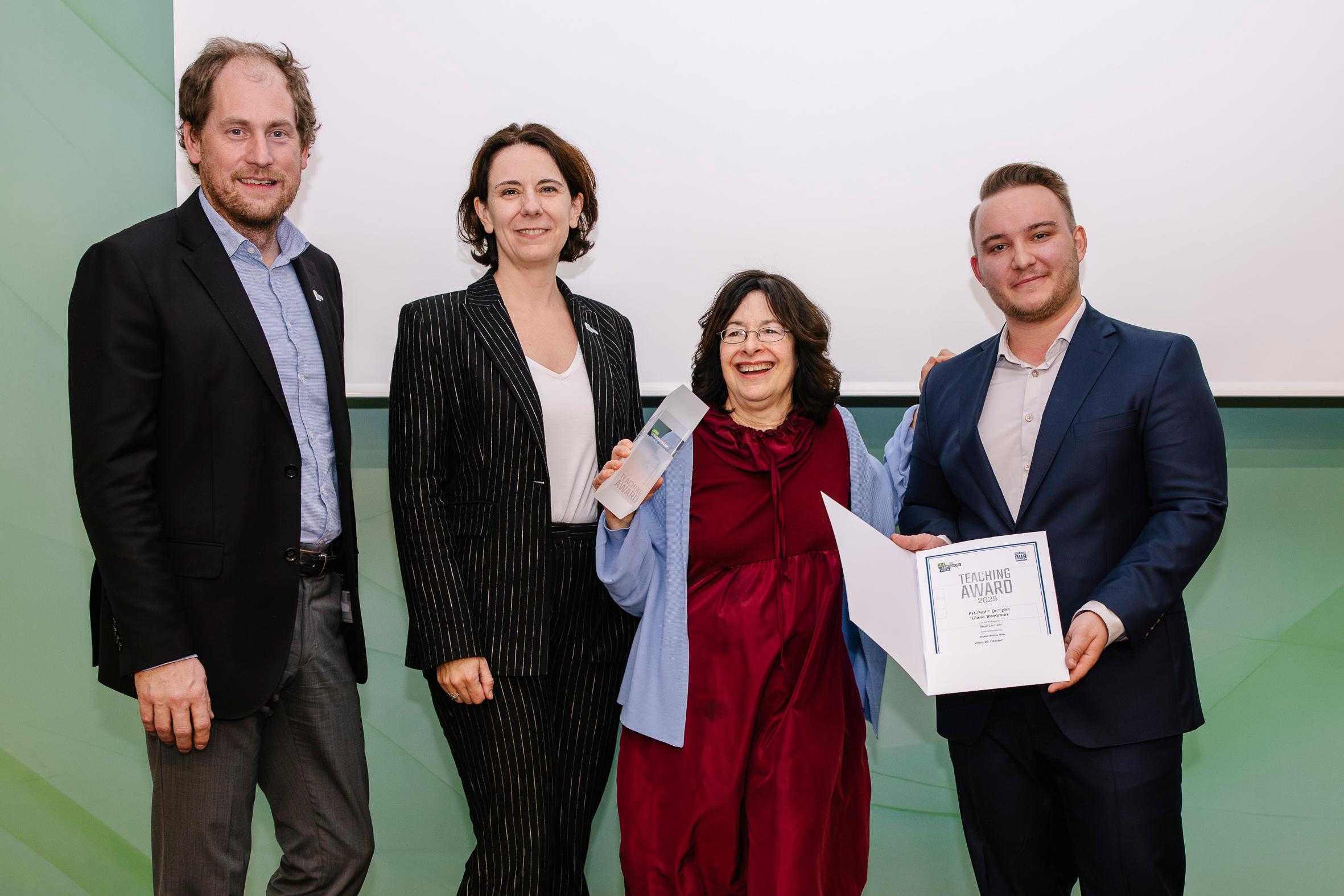 Vier förmlich gekleidete Personen stehen zusammen. Eine Frau hält lächelnd eine Trophäe, während ein anderer Mann eine Urkunde hält. Die Gruppe wirkt glücklich, wahrscheinlich bei einer Preisverleihung, mit einem schlichten Hintergrund hinter ihnen.