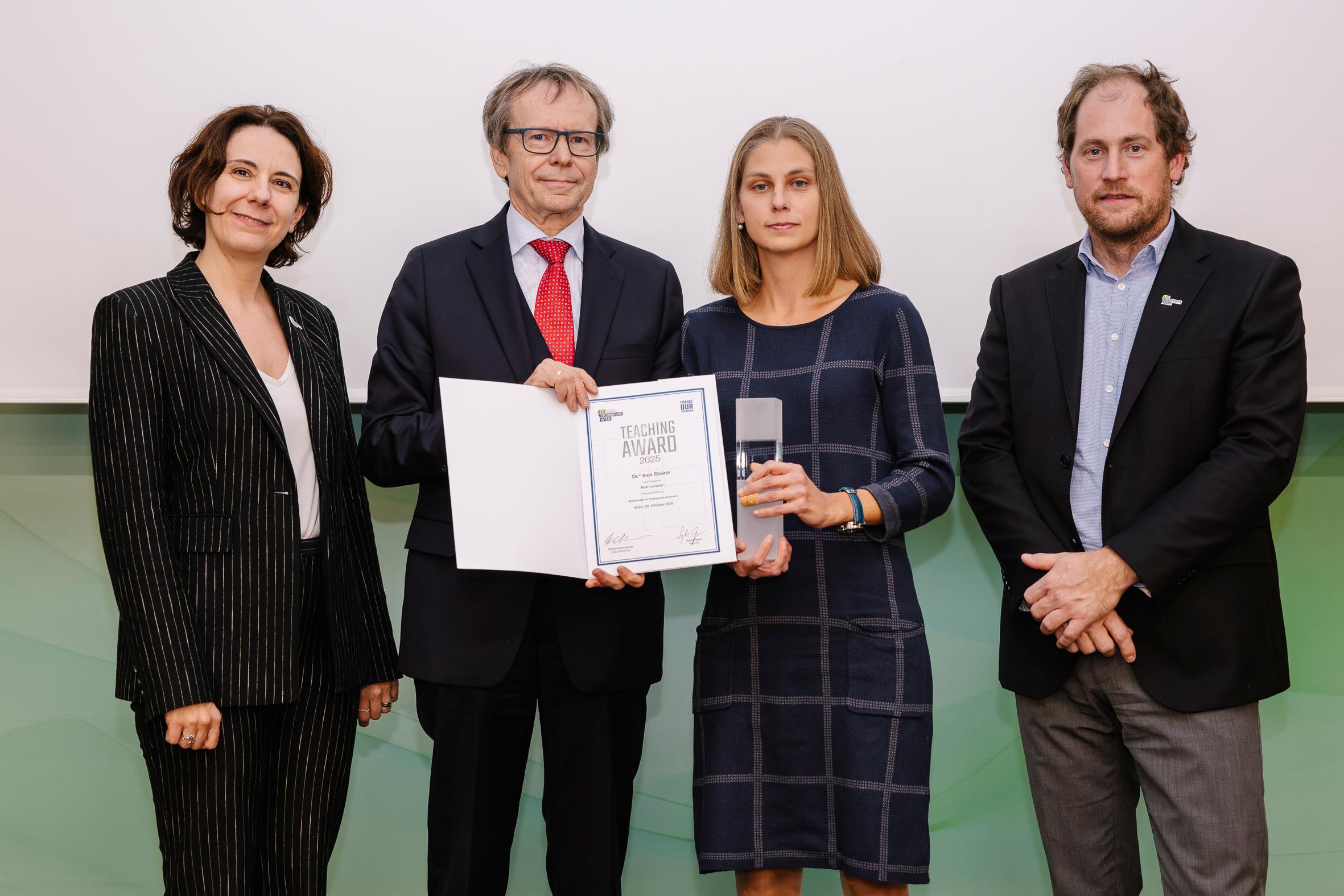 Vier Personen stehen zusammen, während eine Frau eine Urkunde und einen gläsernen Preis in der Hand hält. Sie sind förmlich gekleidet und posieren für ein Foto vor einem schlichten Hintergrund.