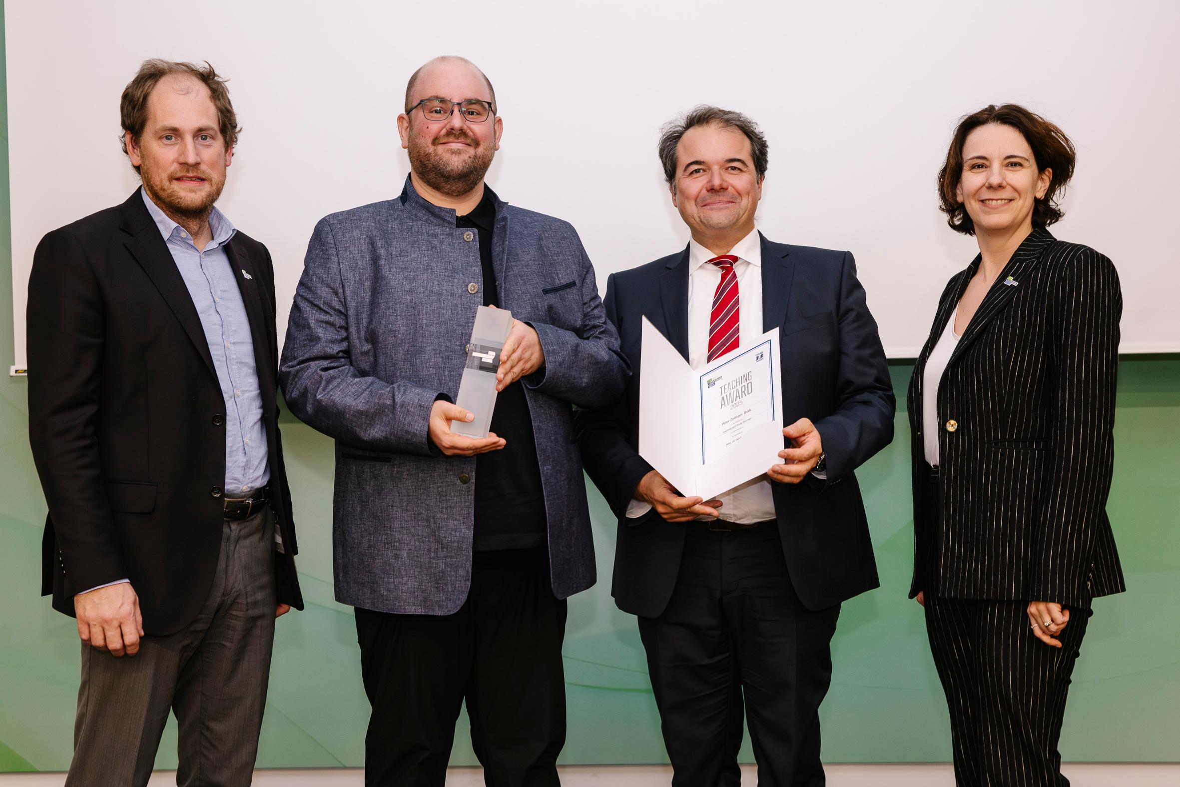 Vier professionell gekleidete Personen stehen lächelnd zusammen. Die zweite Person hält eine Trophäe, und die dritte Person hält eine offene Urkunde oder Auszeichnung. Sie scheinen auf einer formellen Anerkennungs- oder Preisverleihungsveranstaltung zu sein.