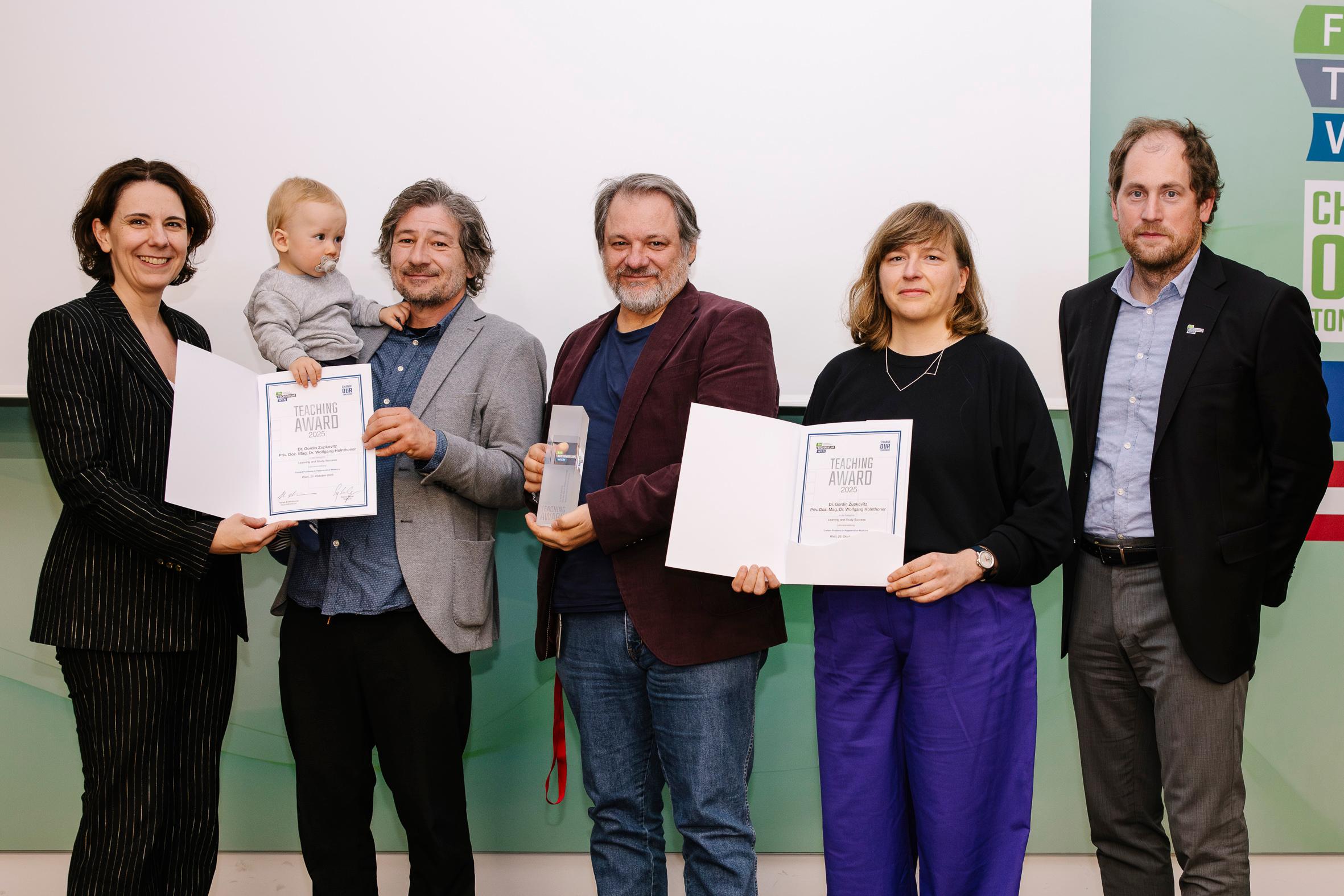 Sechs Erwachsene, von denen einer ein Baby hält, stehen nebeneinander und schauen in die Kamera. Drei von ihnen halten Urkunden und einer eine gläserne Auszeichnung in der Hand. Sie sind halb-formal gekleidet und stehen vor einem weiß-grünen Hintergrund.