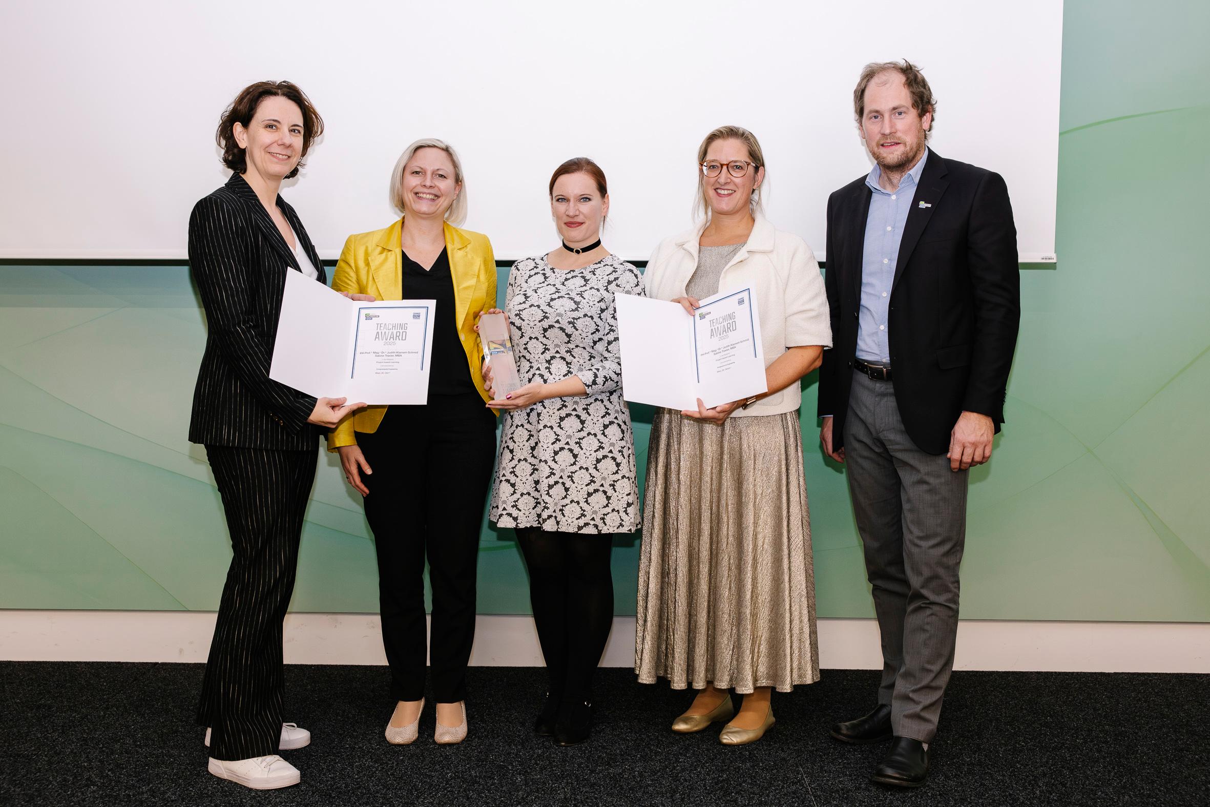 Fünf Personen stehen in einem Innenraum vor einem weiß-grünen Hintergrund; drei Frauen halten Urkunden, eine vierte eine Trophäe. Sie sind formell und halbformell gekleidet und lächeln in die Kamera.