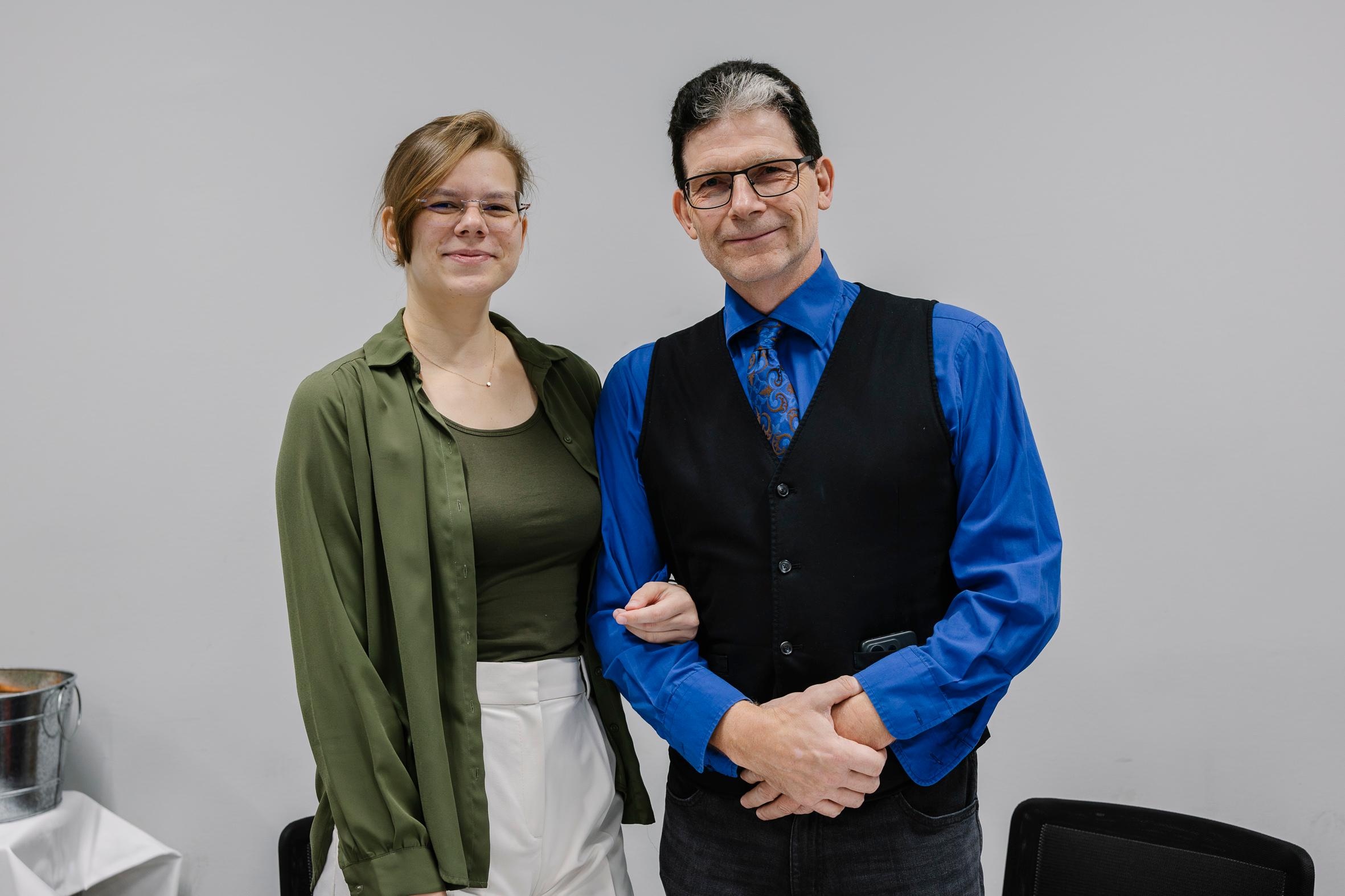 Eine lächelnde Frau in grüner Bluse und weißer Hose steht Arm in Arm mit einem Mann in blauem Hemd, gemusterter Krawatte und schwarzer Weste. Sie stehen drinnen vor einer schlichten grauen Wand, mit Stühlen und einem Tisch in der Nähe.