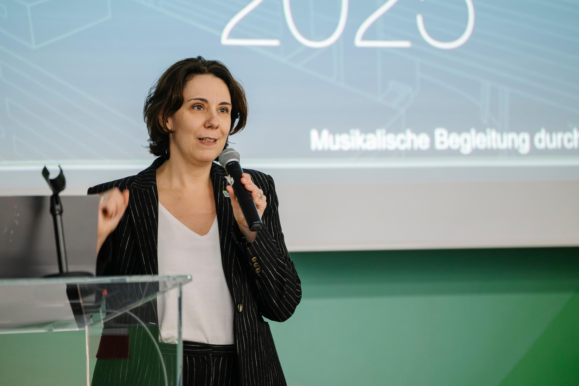 Eine Frau in einem Nadelstreifenanzug spricht in ein Mikrofon, während sie auf einem Podium steht. Hinter ihr ist ein Bildschirm zu sehen, der das Jahr 2023 und einen deutschen Text anzeigt.