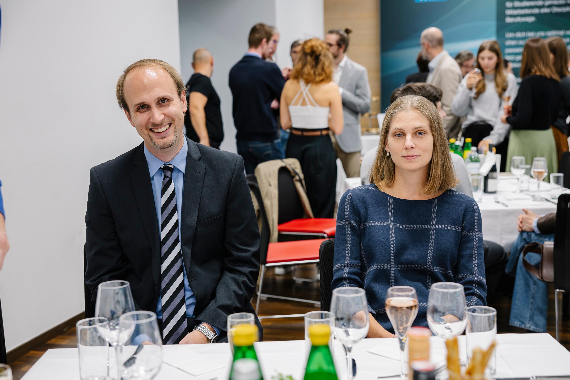 Ein Mann im Anzug und eine Frau im blauen Kleid sitzen an einem mit Gläsern und Getränken gedeckten Tisch und lächeln bei einer Veranstaltung oder Feier, während sich im Hintergrund mehrere Personen unterhalten.