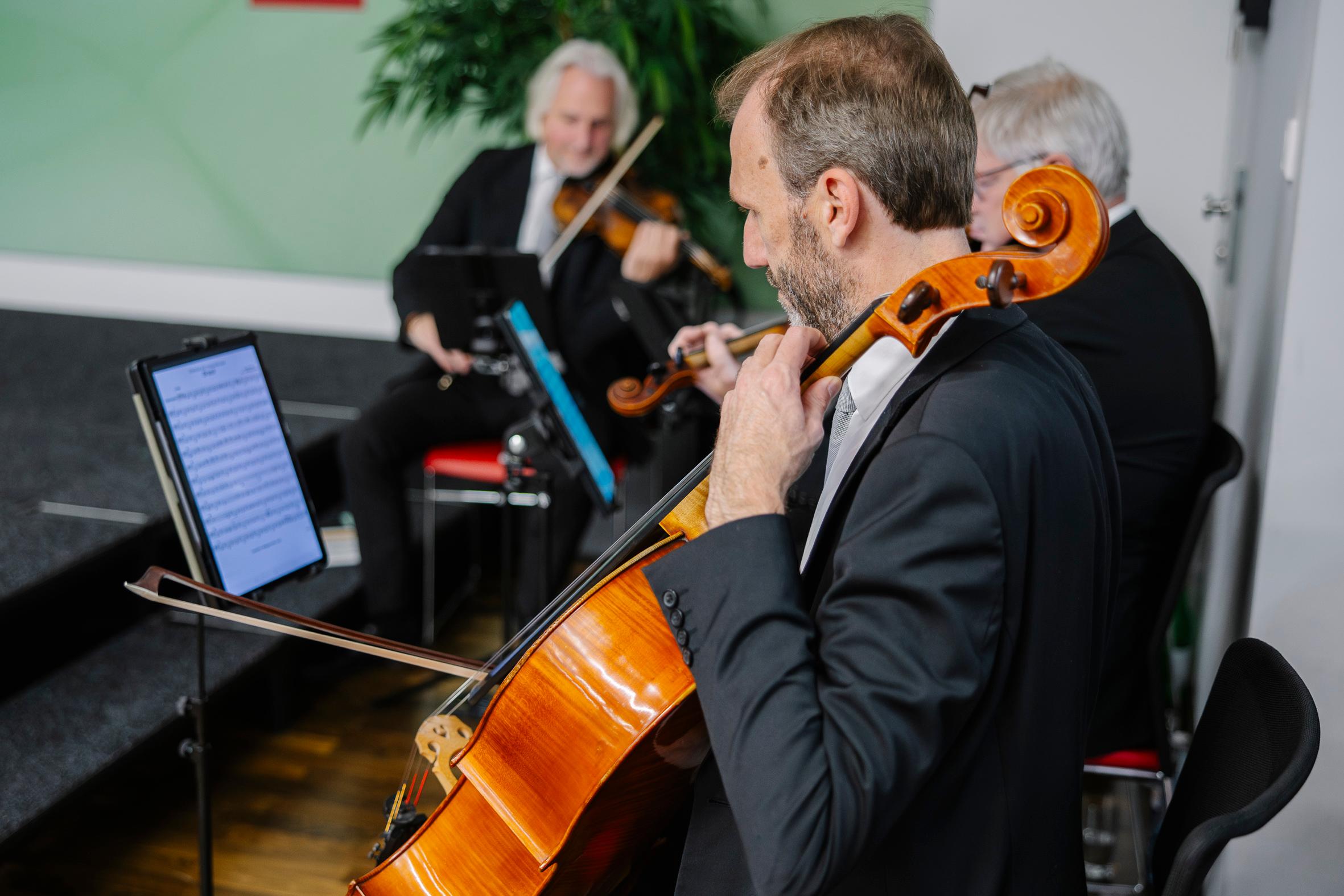 Ein Cellist im Anzug spielt Musik von einem digitalen Tablet, begleitet von zwei Geigern in einem kleinen Ensemble. Die Musiker sitzen in einem Raum und konzentrieren sich auf ihr Spiel.
