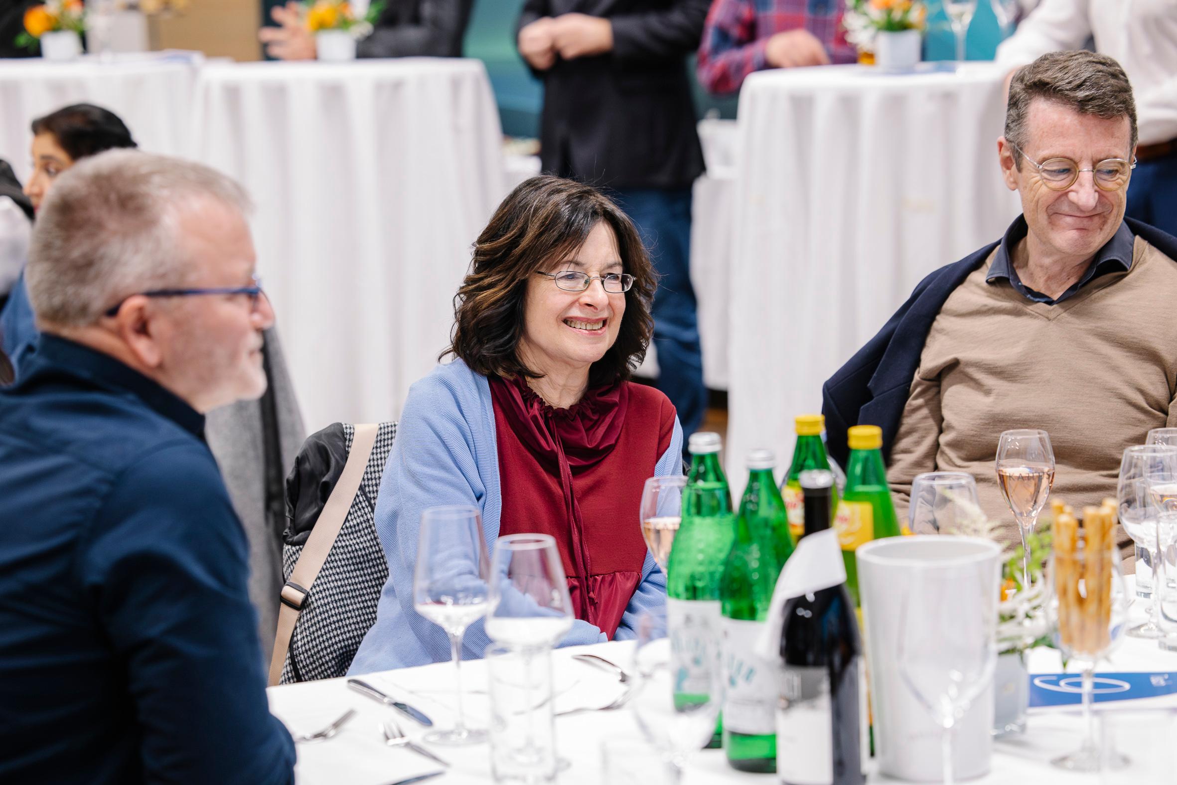 Drei Erwachsene sitzen an einem runden Tisch, der für eine Mahlzeit gedeckt ist, lächeln und unterhalten sich. Auf dem Tisch stehen Wasserflaschen, Weingläser und Besteck. Im Hintergrund sind Menschen und hohe Tische zu sehen, was auf eine gesellschaftliche oder formelle Veranstaltung hindeutet.