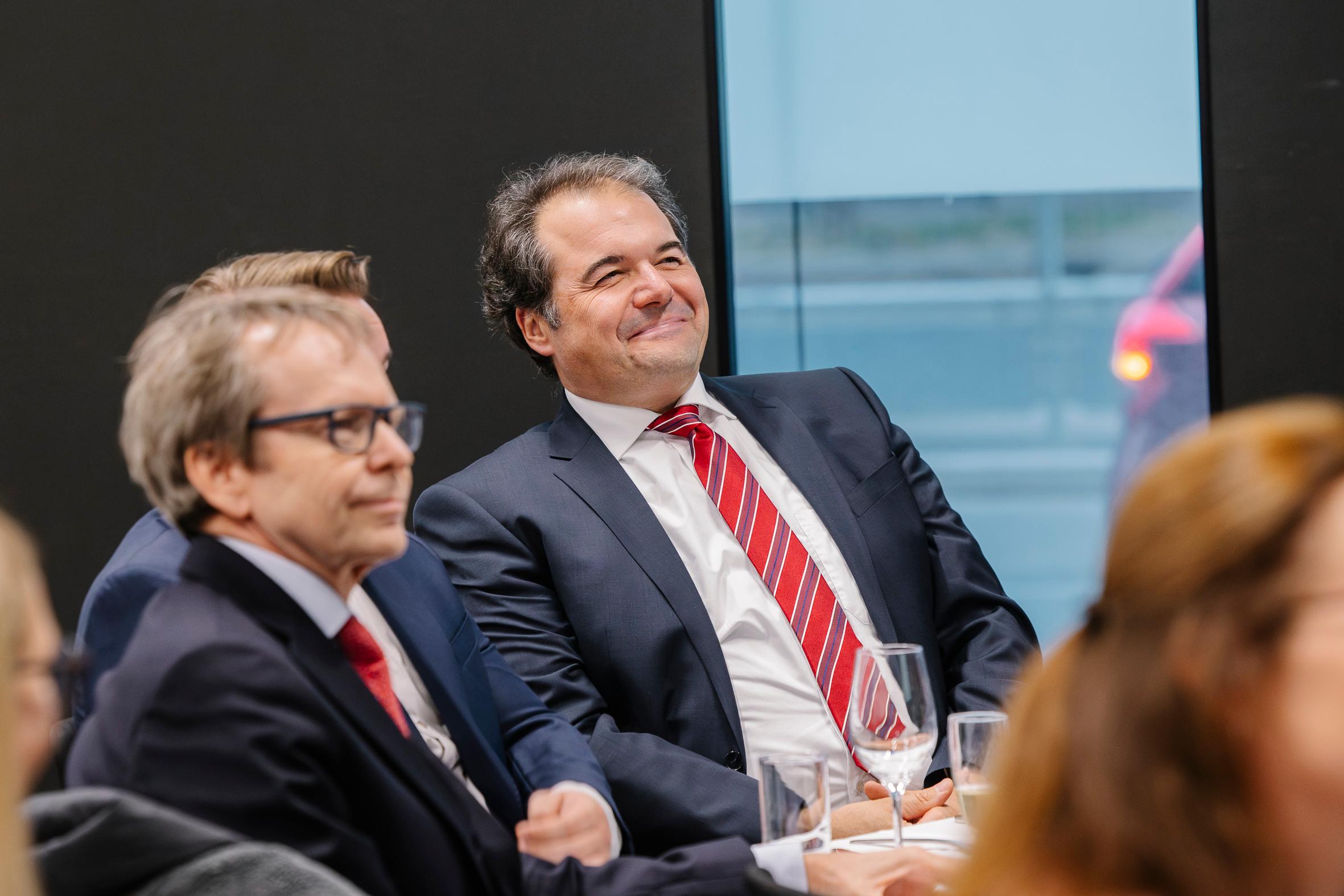 Mehrere Personen in Geschäftskleidung sitzen aufmerksam bei einer Veranstaltung, wobei ein lächelnder Mann im Anzug und mit rot gestreifter Krawatte im Mittelpunkt steht. Auf dem Tisch stehen Weingläser. Im Hintergrund ist ein großes Fenster zu sehen.