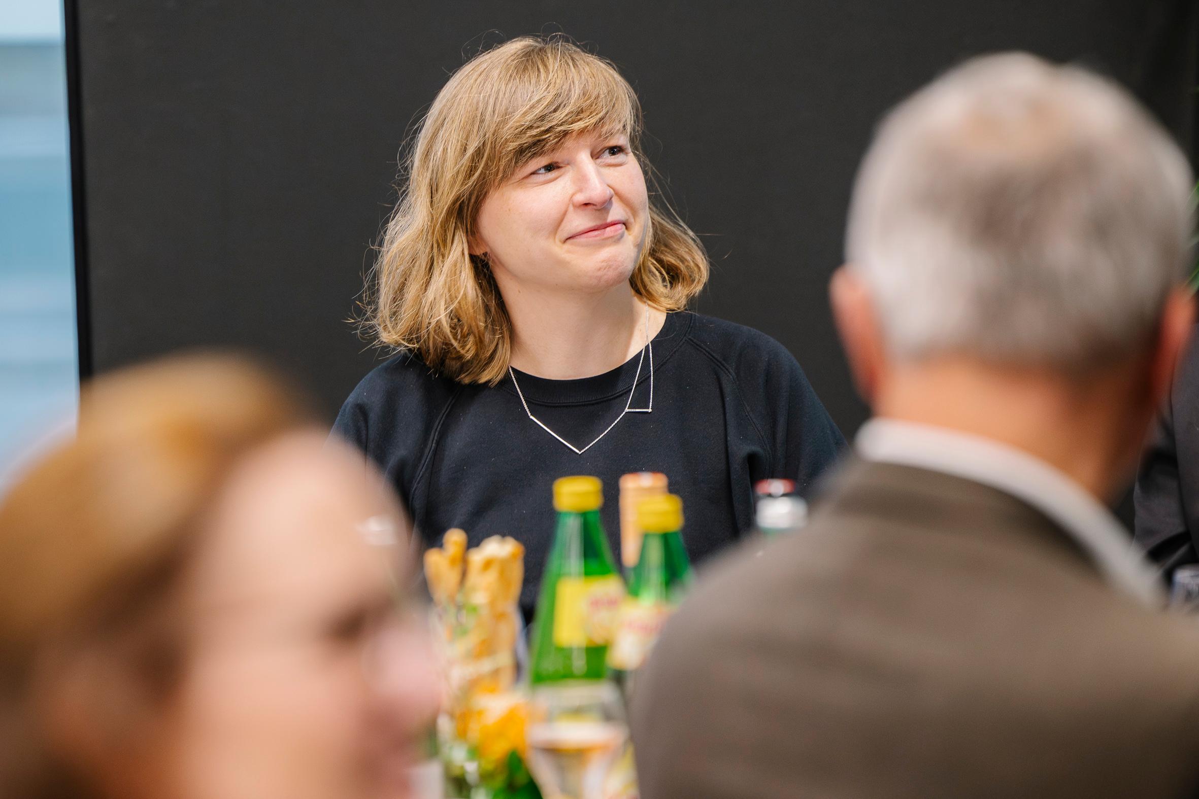 Eine Frau mit schulterlangem blondem Haar sitzt lächelnd an einem Tisch. Auf dem Tisch stehen Wasserflaschen und Gläser, und im Vordergrund sind andere Personen unscharf zu erkennen. Der Hintergrund ist dunkel und einfarbig.