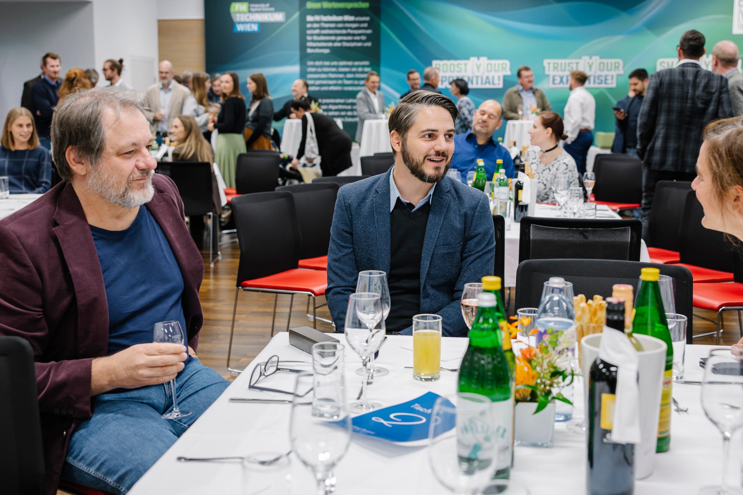 Drei Männer sitzen und unterhalten sich an einem runden Tisch, der für eine formelle Veranstaltung gedeckt ist, mit Getränken und Blumen auf dem Tisch. Im Hintergrund unterhalten sich weitere Personen in der Nähe von großen Plakaten und Bannern an der Wand.