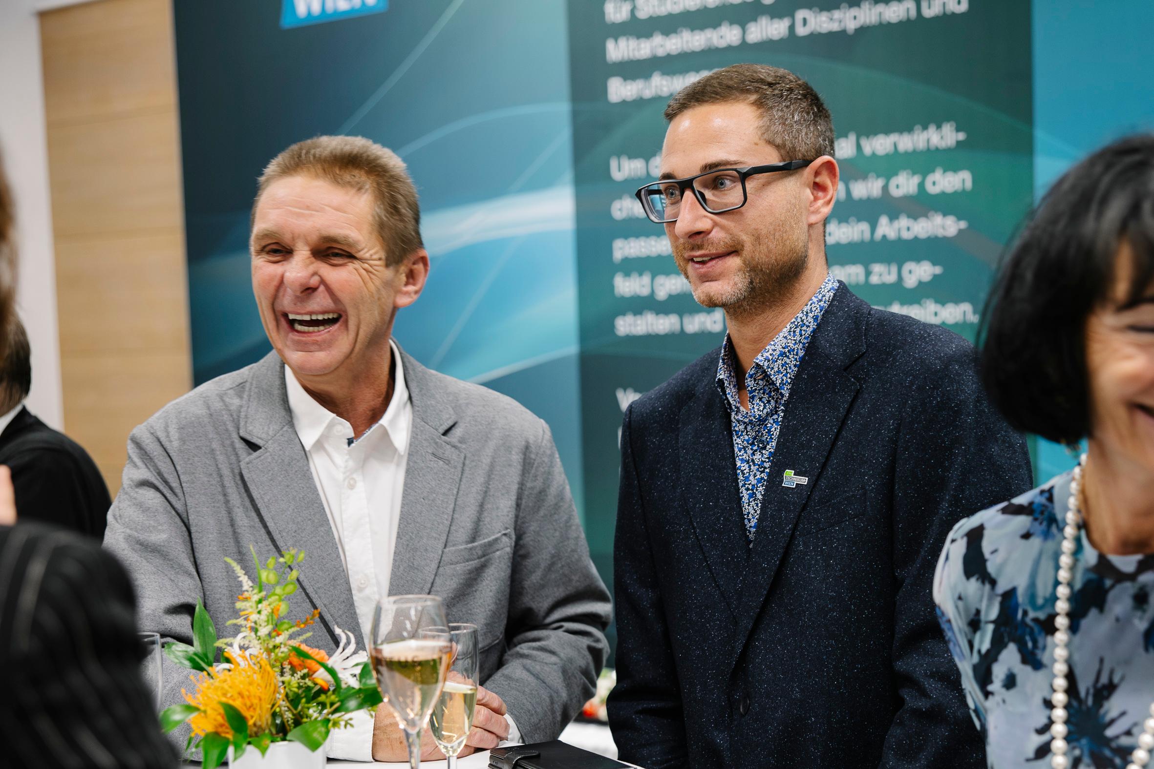 Zwei Männer in Anzügen stehen an einem Tisch mit Getränken und Blumen, lächeln und unterhalten sich bei einer beruflichen Veranstaltung. Eine Frau ist teilweise im Vordergrund zu sehen. Im Hintergrund ist ein blaugrünes Transparent mit deutschem Text zu sehen.