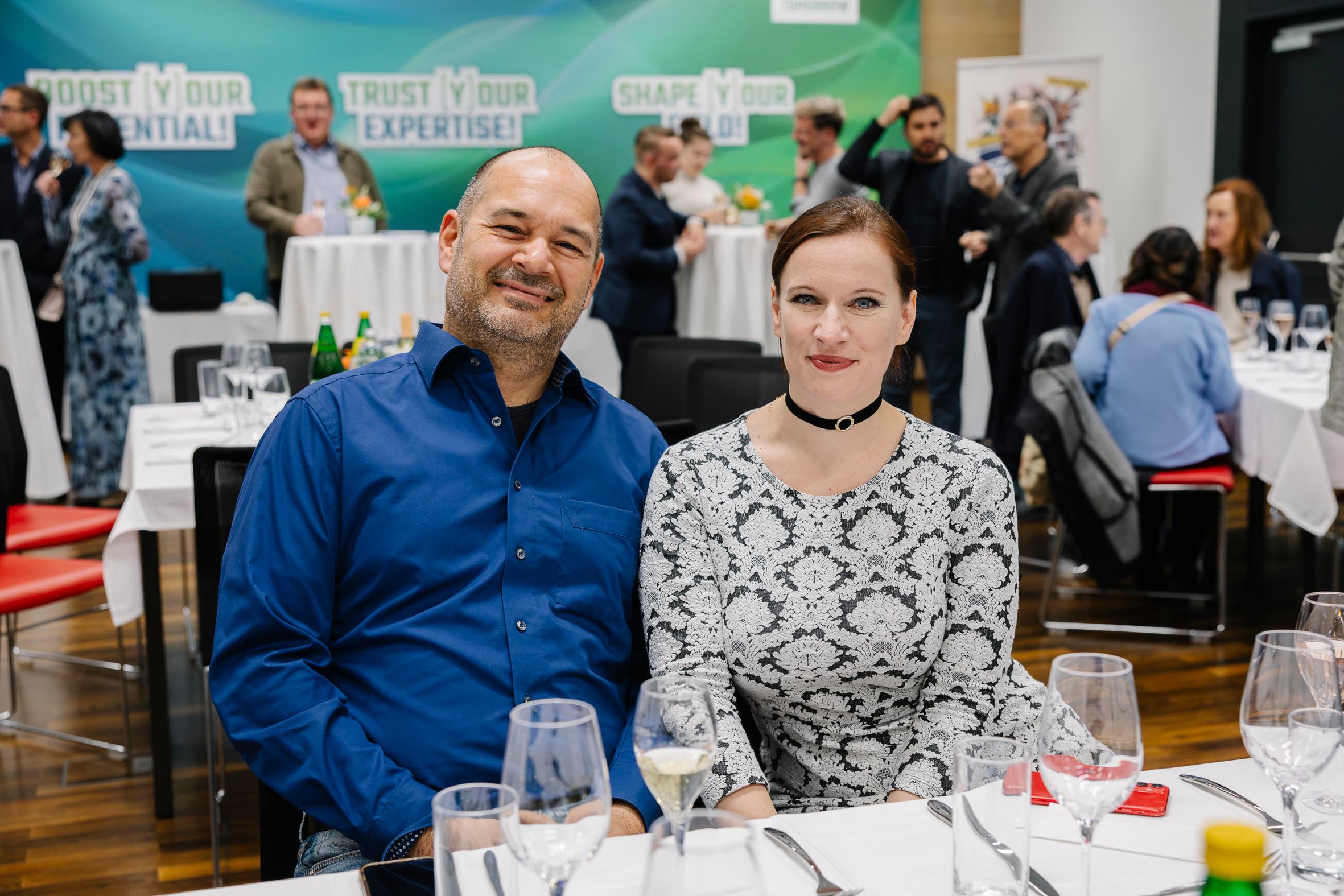 Ein Mann und eine Frau sitzen zusammen an einem Tisch für eine formelle Veranstaltung und lächeln in die Kamera. Hinter ihnen sitzen Menschen in der Nähe von Tischen mit Getränken. Im Hintergrund sind Banner mit motivierendem Text zu sehen.