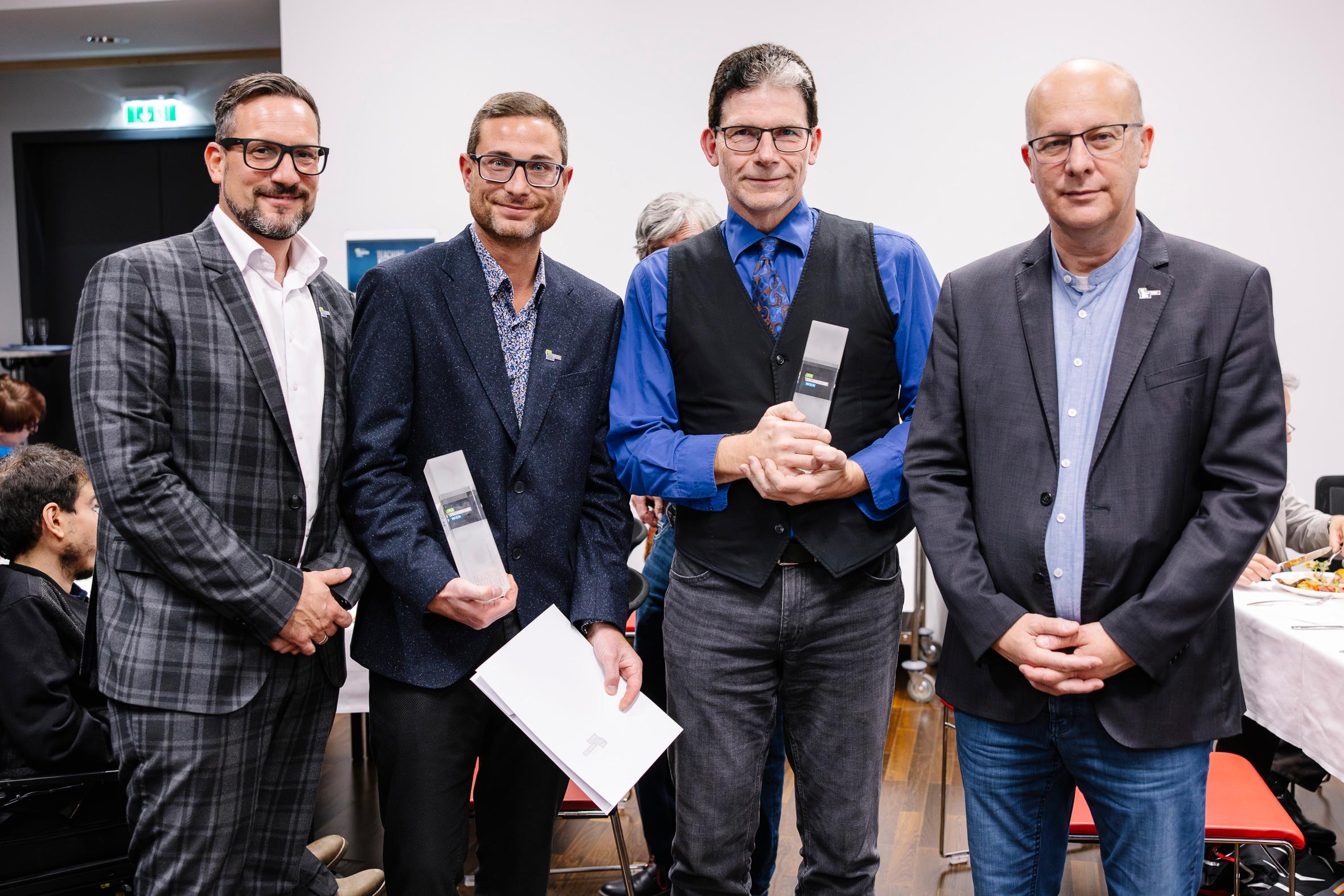 Vier Männer in Geschäftskleidung stehen drinnen, zwei halten gläserne Auszeichnungen und einer hält eine Urkunde. Sie posieren für ein Foto bei einer Preisverleihung oder einer formellen Veranstaltung, wie es scheint. Die Menschen sitzen an Tischen im Hintergrund.