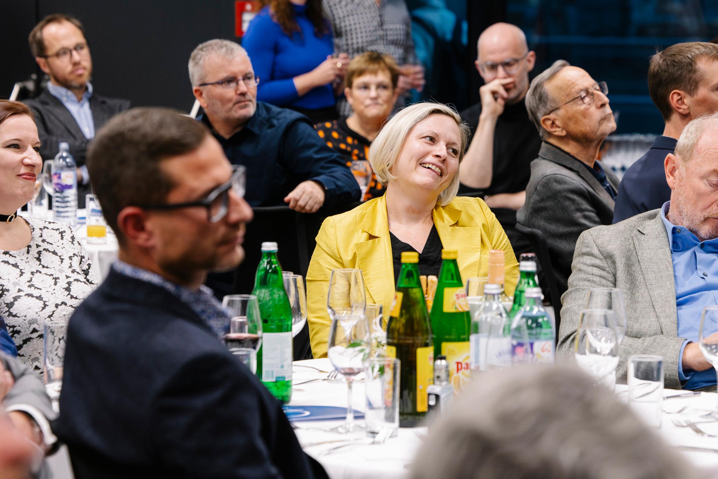 Eine Gruppe von Menschen sitzt an runden Tischen mit Getränken und hört bei einer Veranstaltung in einer Halle aufmerksam zu. Eine Frau in einer gelben Jacke lächelt in der Mitte, während andere zu einem Redner außerhalb des Blickfelds schauen.