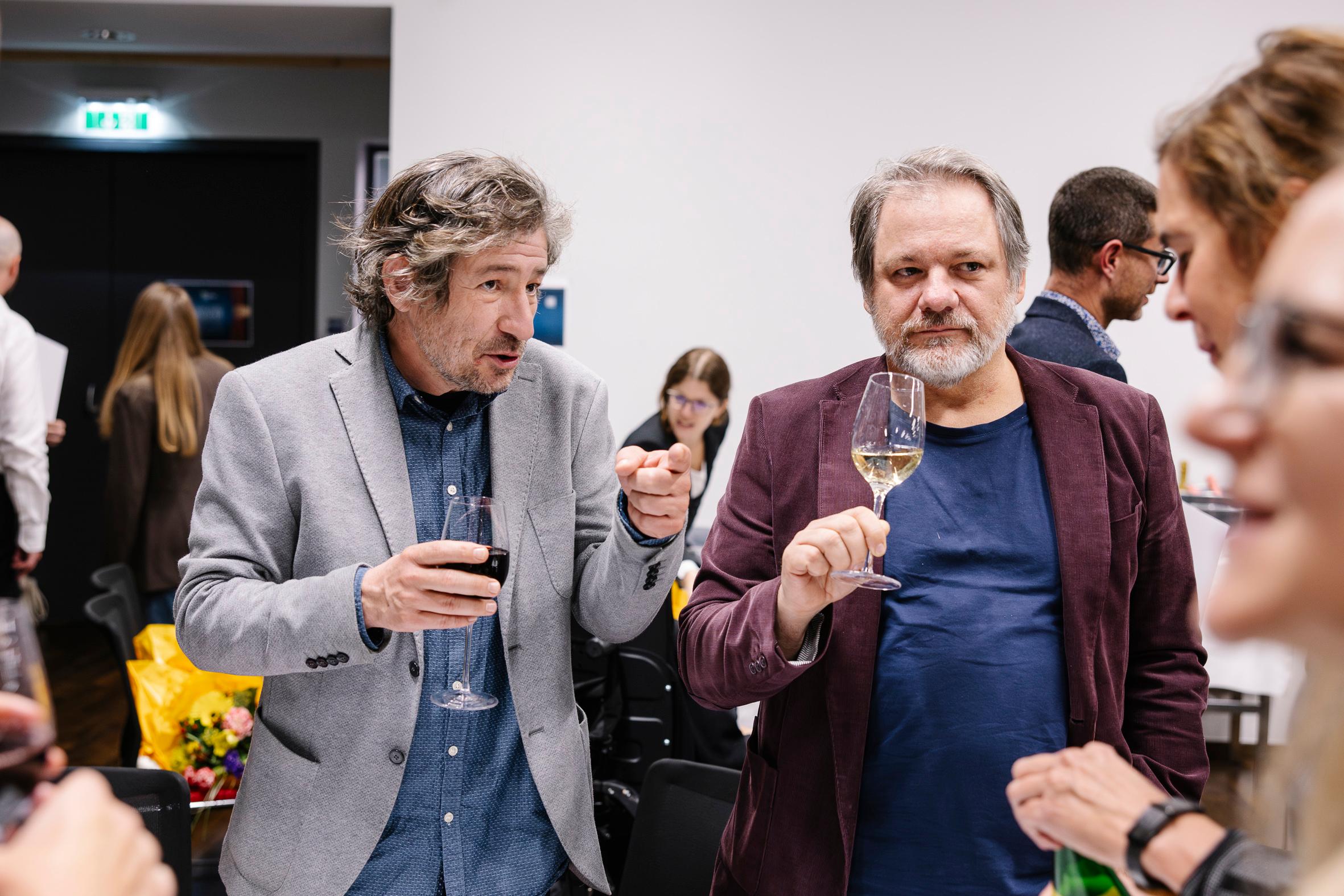 Zwei Männer stehen bei einem gesellschaftlichen Ereignis zusammen, jeder hält ein Glas Wein in der Hand und unterhält sich. Andere Leute mischen sich im Hintergrund in einem hell erleuchteten Raum.