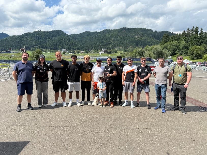 Eine Gruppe von Menschen, darunter Erwachsene und ein Kind, steht Seite an Seite im Freien auf einem Bürgersteig vor einer Kulisse aus grünen Hügeln, Bäumen und einem bewölkten Himmel. Einige tragen schwarze Hemden, und alle lächeln in die Kamera.