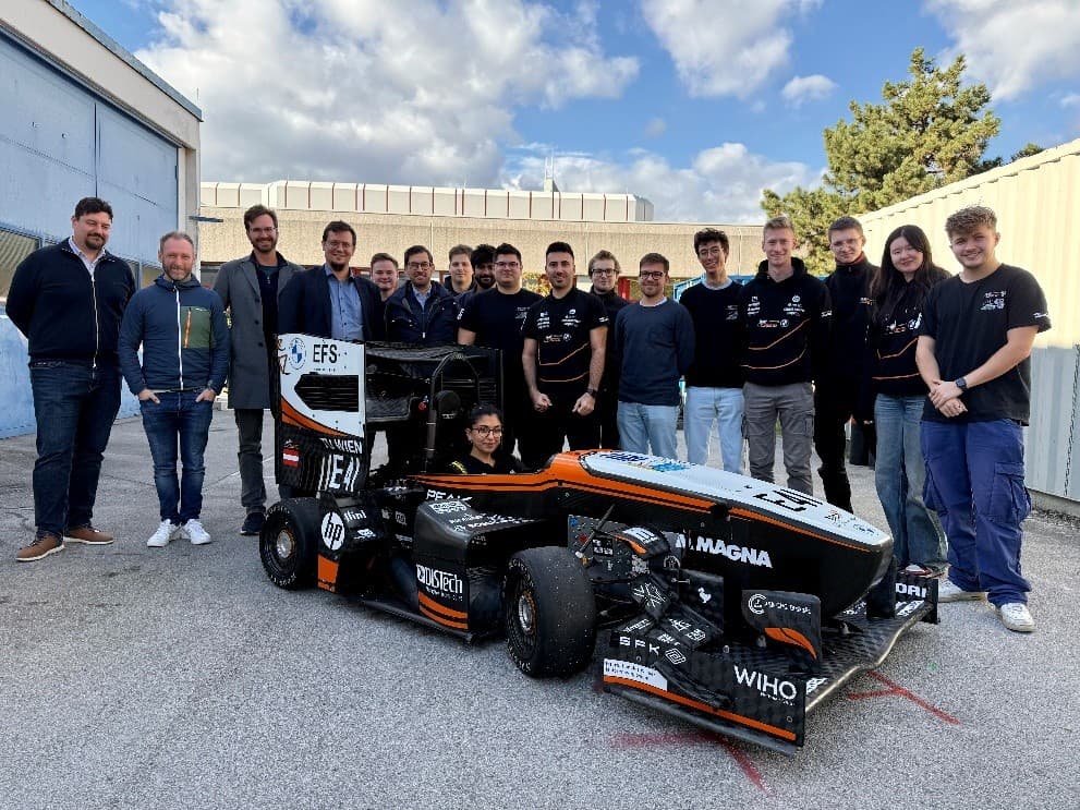 Eine Gruppe von Menschen steht lächelnd um einen kleinen schwarz-orangen Rennwagen, der im Freien auf einer gepflasterten Fläche mit Gebäuden und einem Baum im Hintergrund geparkt ist. Eine Person sitzt im Inneren des Wagens.