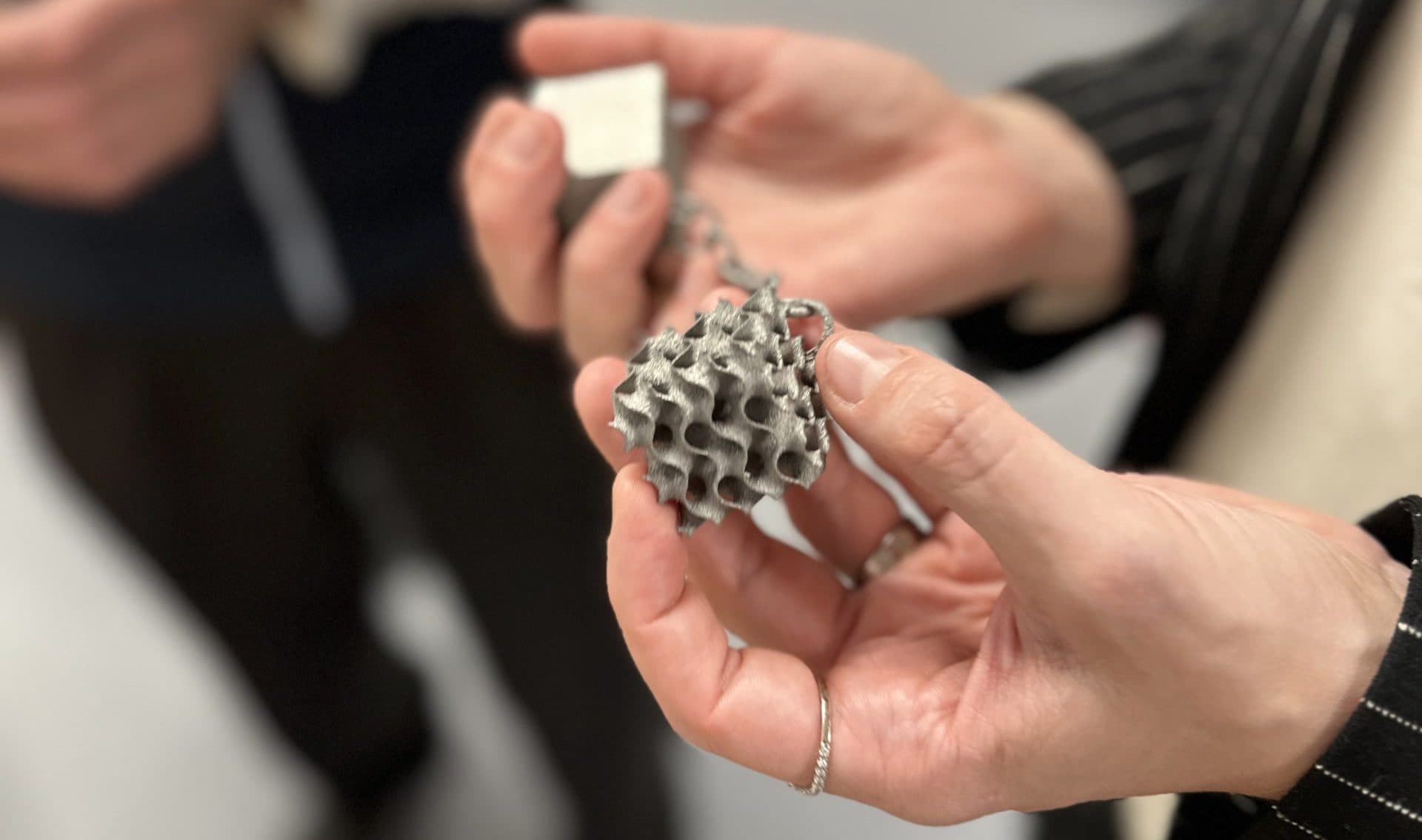Eine Person hält einen 3D-gedruckten Metallgegenstand mit einer wabenartigen Struktur, während eine andere Person in der Nähe einen kleinen Würfel hält; beide sind in einem Raum und tragen Geschäftskleidung.