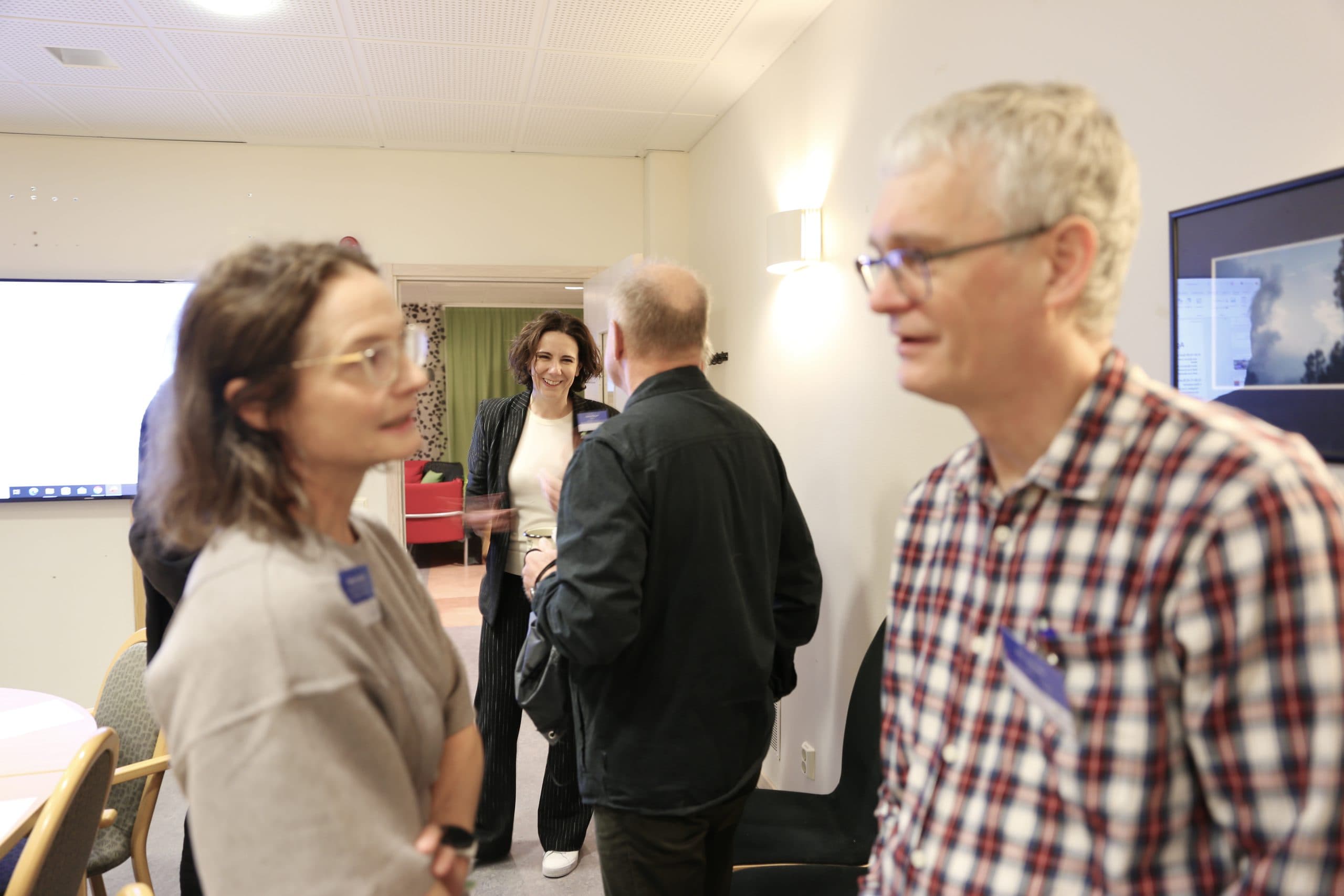 Vier Erwachsene unterhalten sich in einem gut beleuchteten Raum. Zwei Personen im Vordergrund unterhalten sich, eine trägt ein kariertes Hemd und eine Brille. Im Hintergrund lächelt eine Frau eine andere Person an, die mit dem Rücken zur Kamera steht.