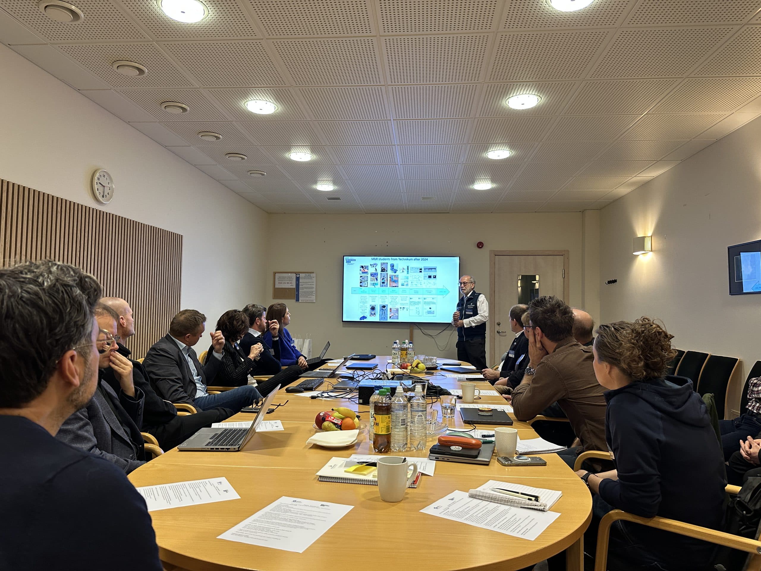 Eine Gruppe von Personen sitzt um einen Konferenztisch und hört einer Person zu, die Folien auf einem Bildschirm im vorderen Teil des Raums präsentiert. Auf dem Tisch stehen Papiere, Laptops, Getränke und Snacks. Der Raum ist mit hellen Deckenleuchten ausgestattet.