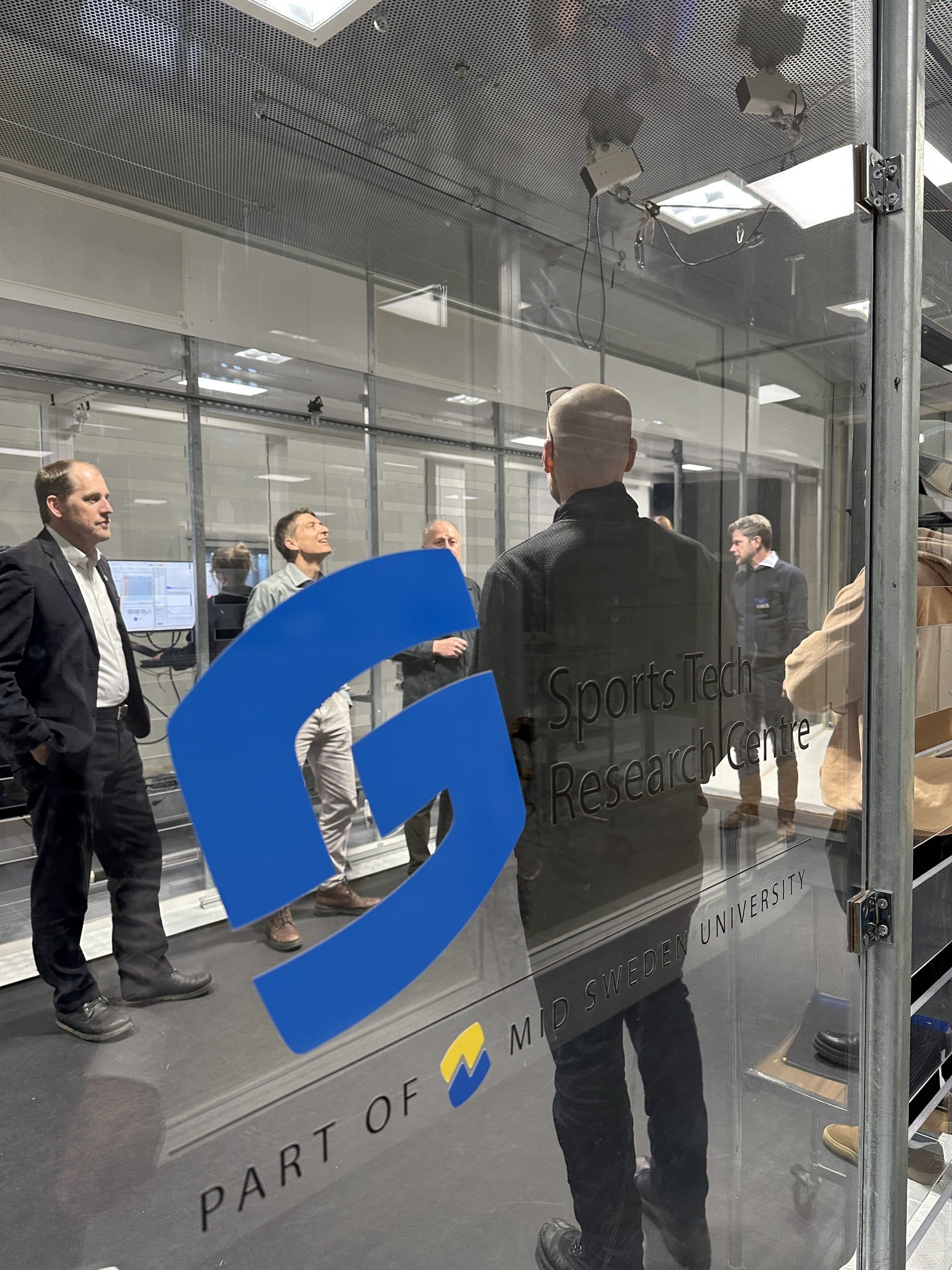Eine Gruppe von Menschen steht und unterhält sich in einem modernen Labor des Sports Tech Research Centre, das Teil der Mid Sweden University ist. Die Szene wird durch eine Glaswand betrachtet, auf der das blaue Logo und der Name des Zentrums zu sehen sind.