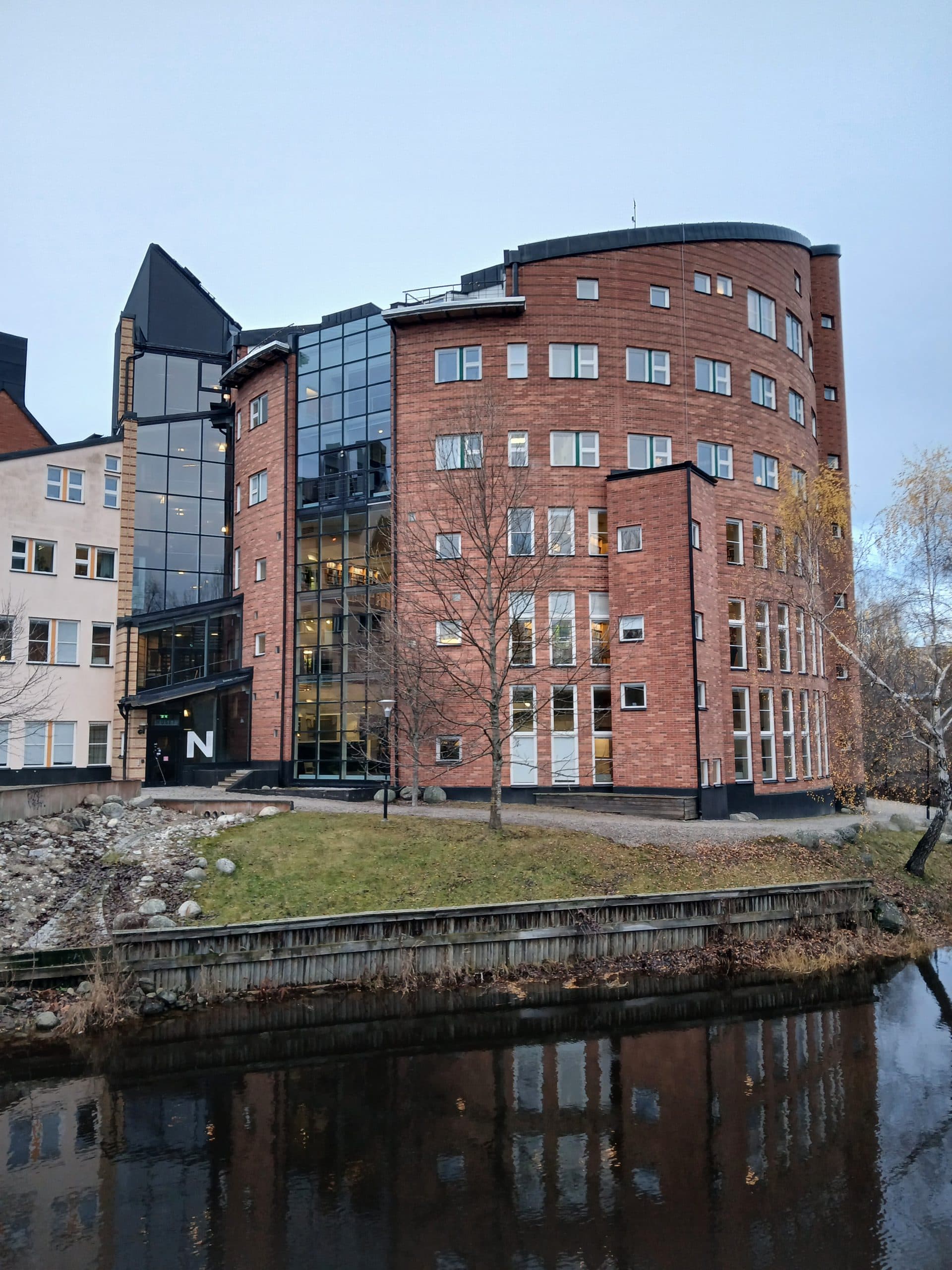 Ein modernes, mehrstöckiges Bürogebäude aus rotem Backstein mit großen Glasfenstern steht an einem Flussufer, das sich im ruhigen Wasser spiegelt. Im Vordergrund sind blattlose Bäume und eine Grasfläche zu sehen, über denen sich ein bewölkter Himmel spiegelt.