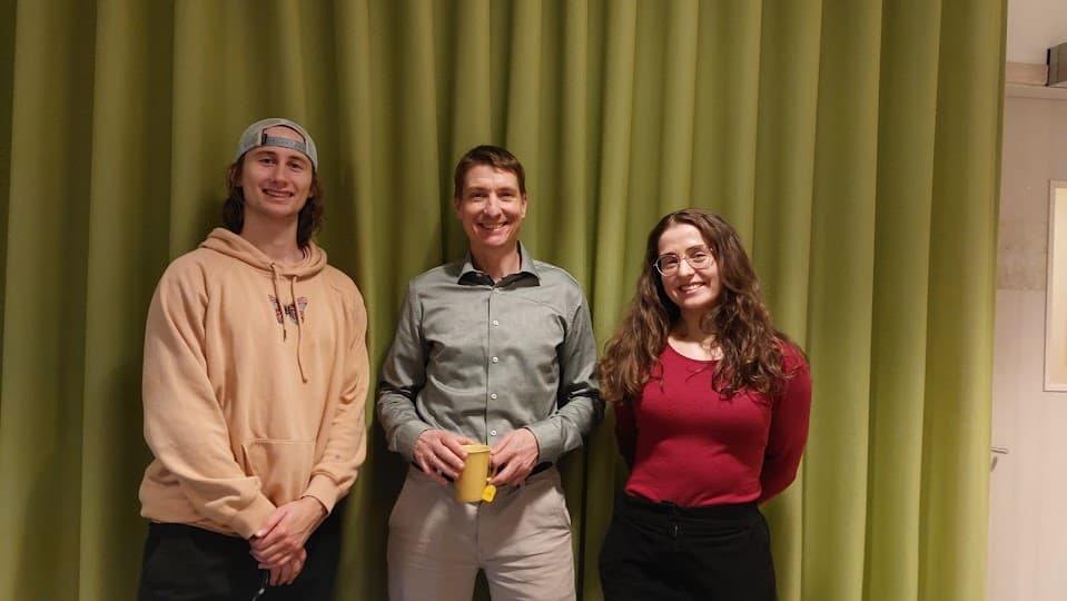 Drei Personen stehen vor einem grünen Vorhang und lächeln in die Kamera. Die Person links trägt einen beigen Kapuzenpulli und eine umgekehrte Mütze, die Person in der Mitte ein graues Hemd mit einem gelben Becher in der Hand, und die Person rechts trägt eine Brille und ein rotes Oberteil.