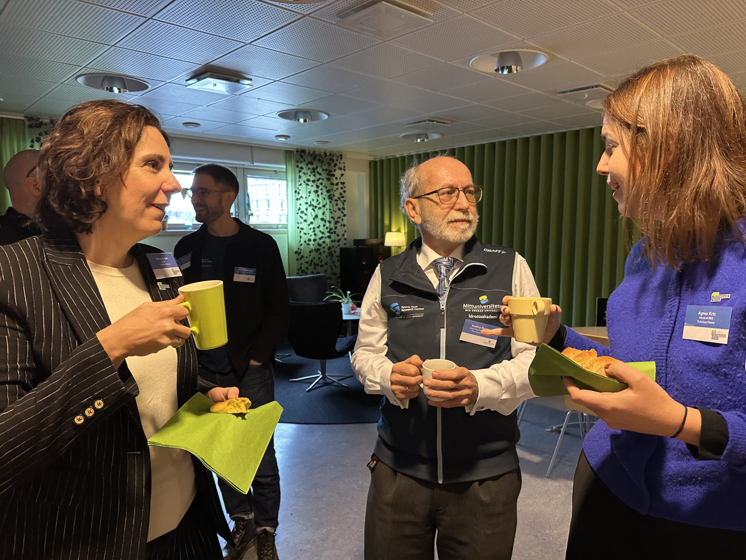 Drei Personen stehen in einem Innenraum zusammen, halten Tassen und grüne Servietten in der Hand und unterhalten sich bei einer geschäftlichen Veranstaltung. Zwei Frauen und ein Mann sind zu sehen, alle tragen Namensschilder und Geschäftskleidung. Andere sind unscharf im Hintergrund zu erkennen.