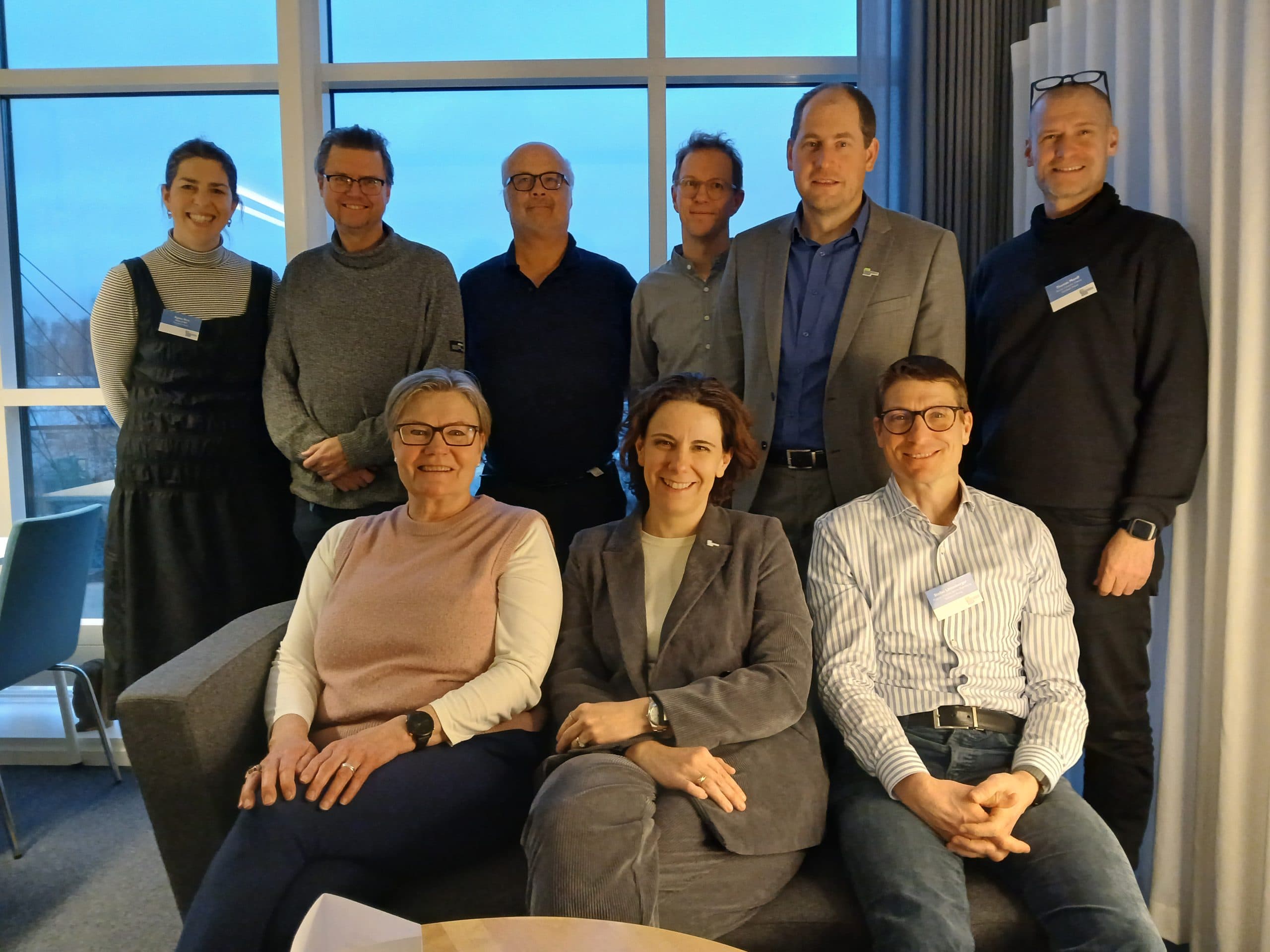 Neun Erwachsene, sechs stehend und drei sitzend auf einer Couch, posieren und lächeln für ein Gruppenfoto in einem Raum mit großen Fenstern und Vorhängen. Die Umgebung wirkt leger und professionell.