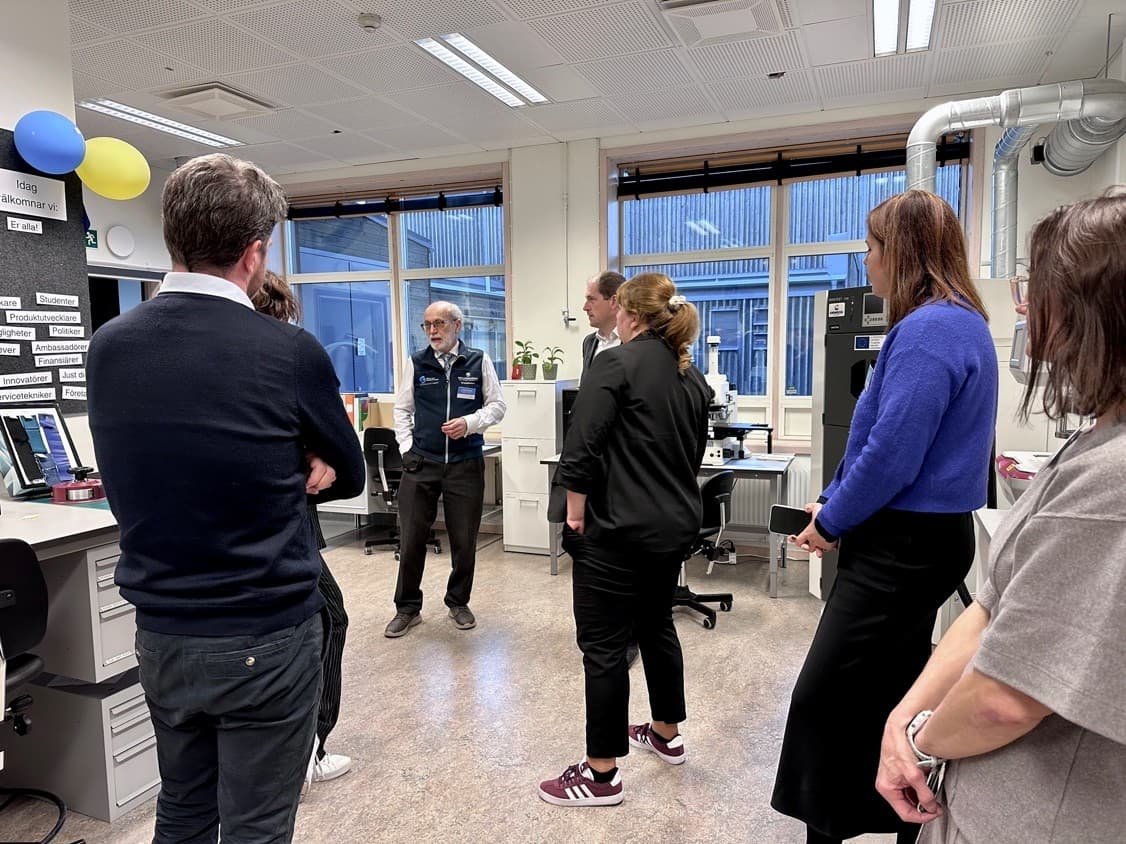 Eine Gruppe von Menschen steht in einem Büro oder Labor und hört einem älteren Mann zu, der spricht. In dem Raum sind Schreibtische, Computer, Ballons und verschiedene Geräte zu sehen. Große Fenster lassen natürliches Licht herein.