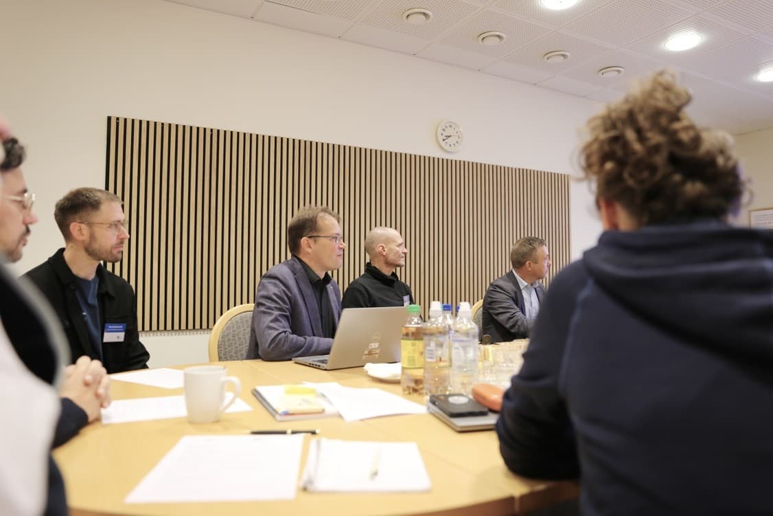 Eine Gruppe von fünf Personen sitzt um einen Konferenztisch und hört aufmerksam zu. Auf dem Tisch befinden sich Laptops, Notebooks und Wasserflaschen. Die Umgebung ist ein heller Besprechungsraum mit einer Uhr und einer Holztafel an der Wand.