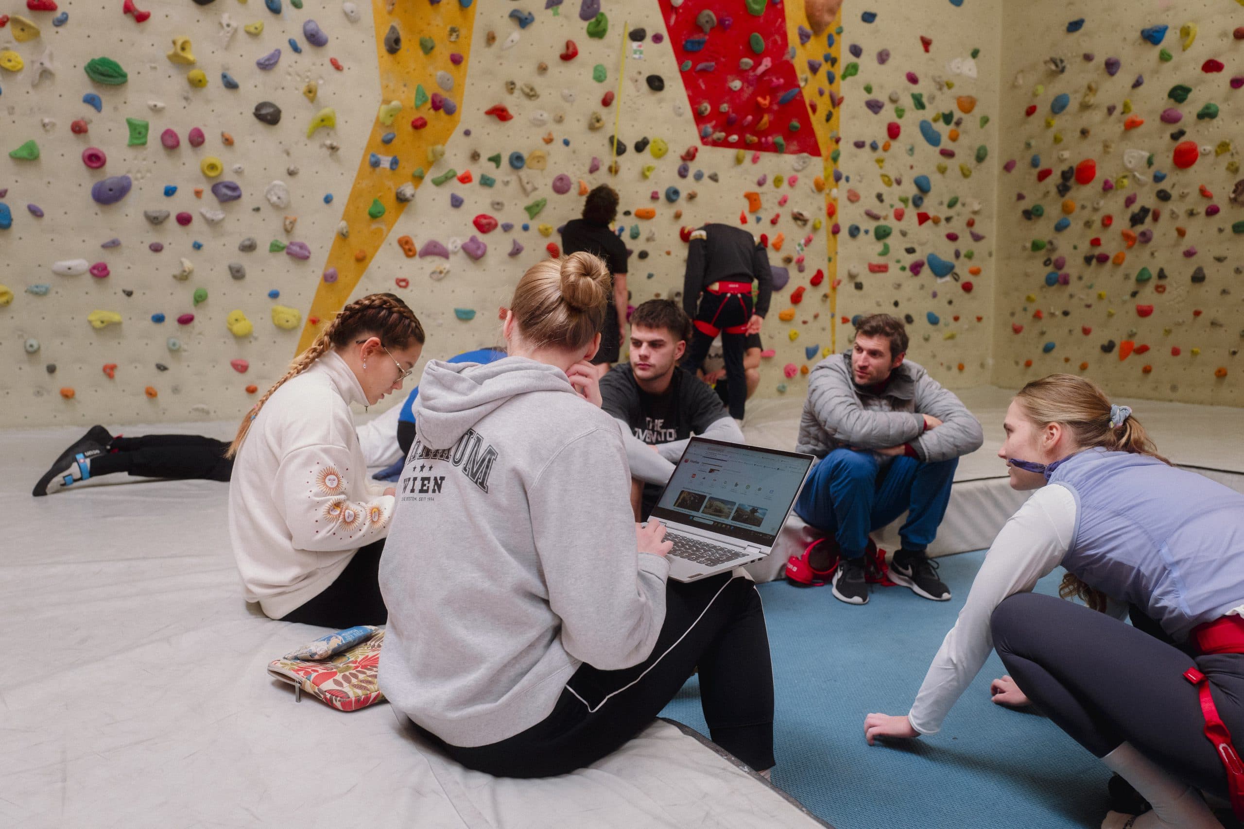 Eine Gruppe junger Leute sitzt auf dem Boden einer Kletterhalle und unterhält sich, während hinter ihnen bunte Klettergriffe an den Wänden hängen. Eine Person arbeitet an einem Laptop, andere dehnen sich oder unterhalten sich.