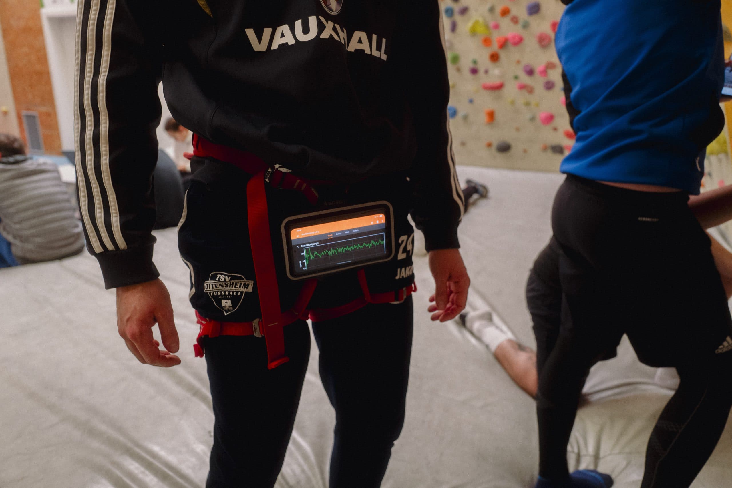Eine Person, die einen Klettergurt trägt, an dessen Taille ein Gerät mit einem Herzfrequenzmesser befestigt ist, steht in einer Kletterhalle. Im Hintergrund sind andere Personen zu sehen.