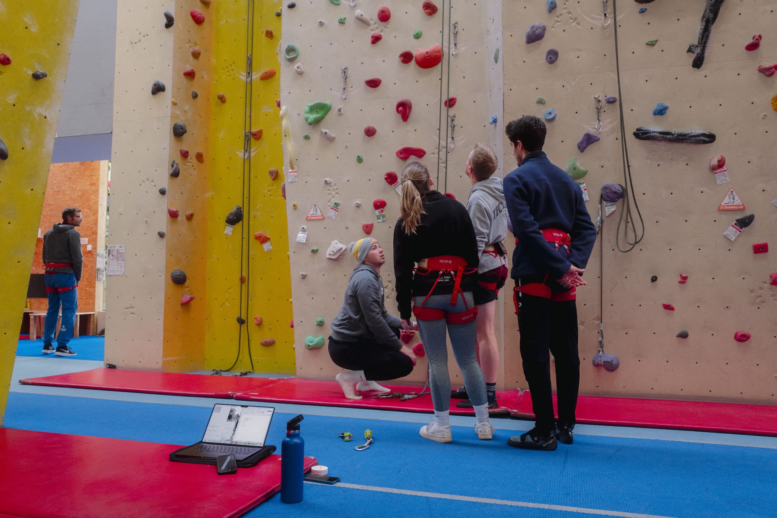 Vier Personen in Klettergurten stehen an einer Indoor-Kletterwand und hören einem Ausbilder zu, der sich hinkniet und etwas erklärt. In der Nähe liegen ein Laptop und eine Wasserflasche auf einer roten Matte. Eine weitere Person steht im Hintergrund.