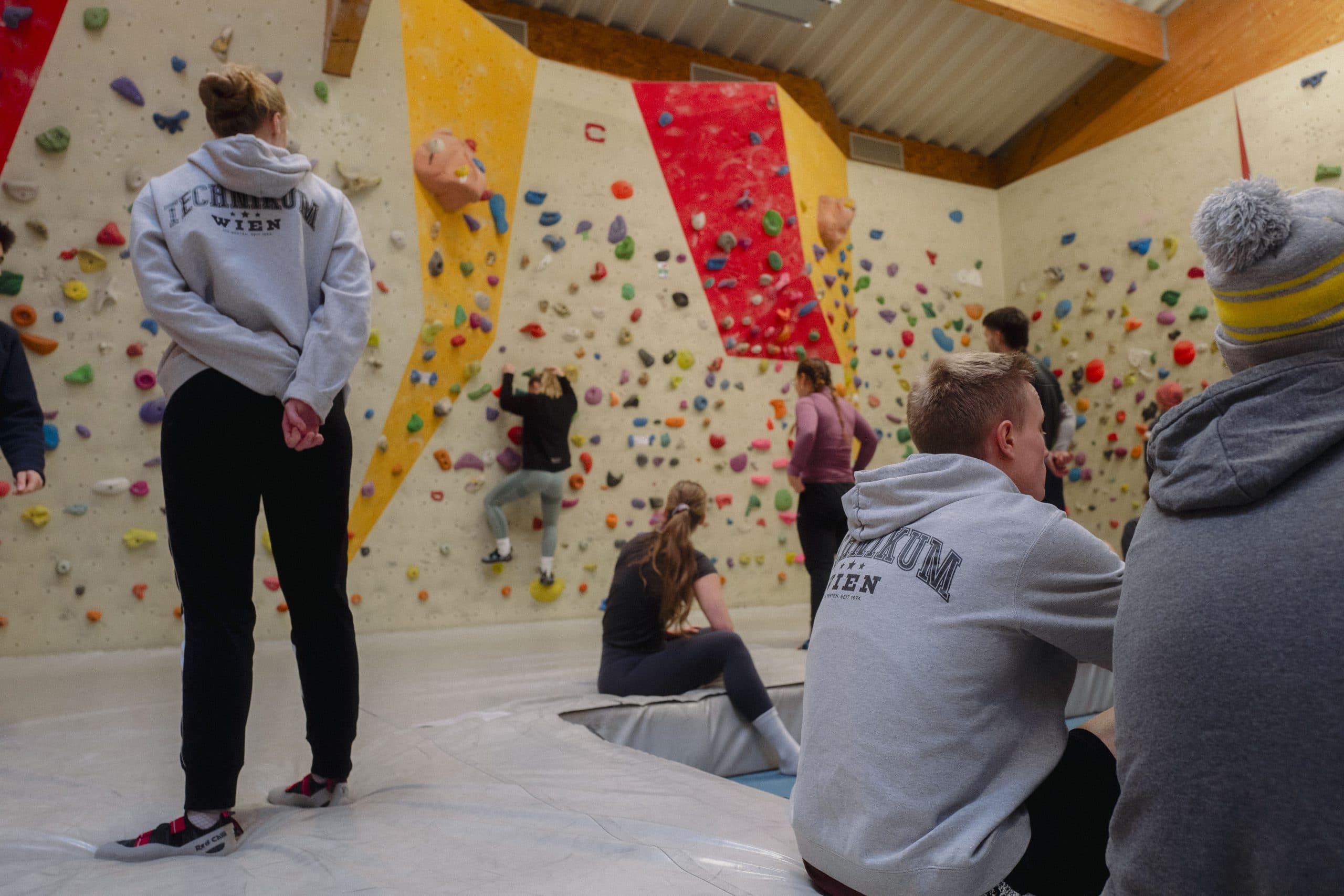 Menschen in Sportkleidung bouldern und schauen anderen beim Klettern an einer Indoor-Kletterwand mit bunten Griffen zu. Einige sitzen auf gepolsterten Matten, während andere stehen oder sich zum Klettern vorbereiten.